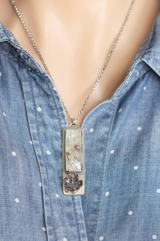 A sterling silver necklace with a sugar water flower agate pendant and succulent accent, worn on a model against a blue polka dot shirt.