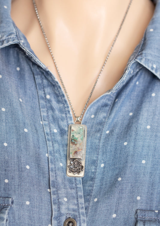 A sterling silver necklace with a sugar water flower agate pendant and succulent accent, worn on a model against a blue polka dot shirt.