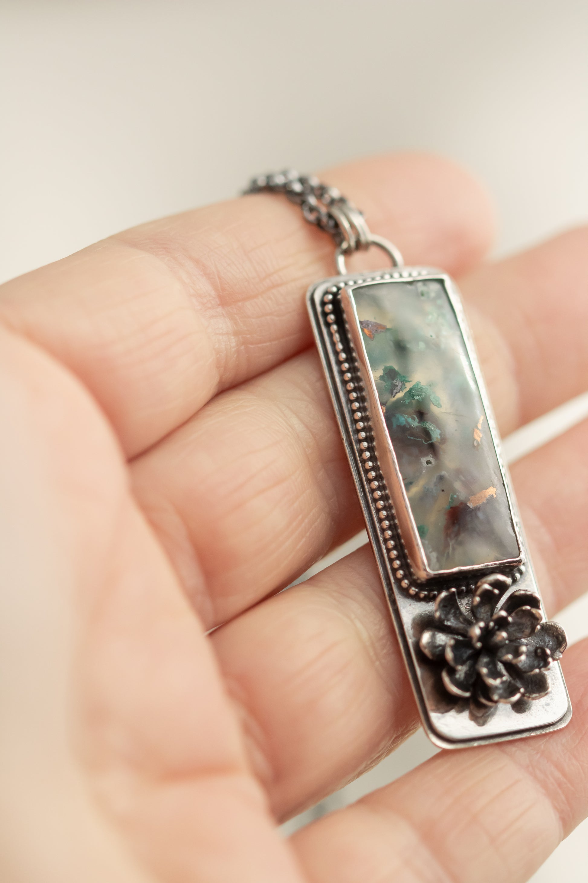 : Close-up of a sterling silver sugar water flower agate necklace with a succulent accent, held in hand, showcasing the pendant's details.