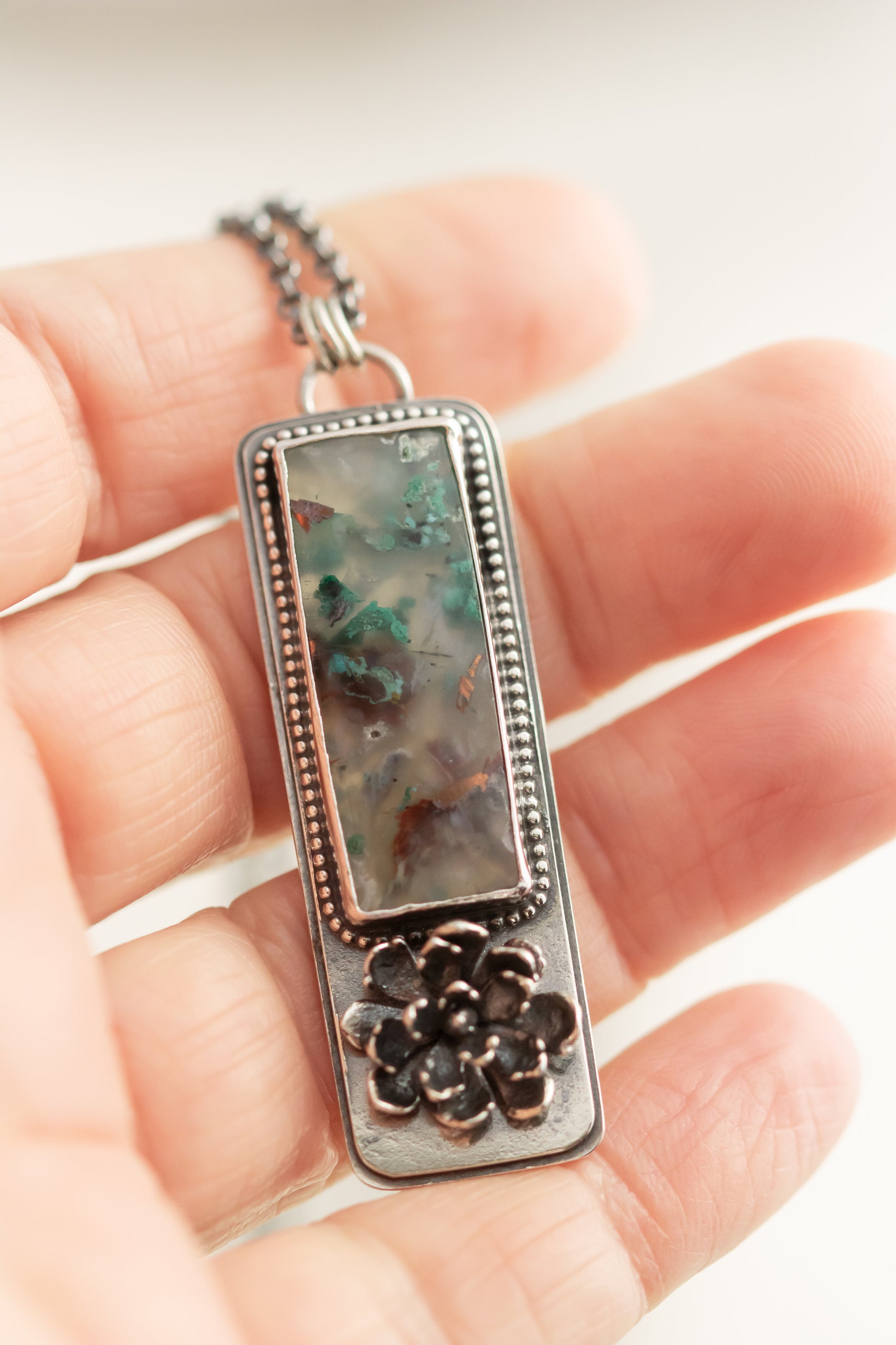 : Close-up of a sterling silver sugar water flower agate necklace with a succulent accent, held in hand, showcasing the pendant's details.
