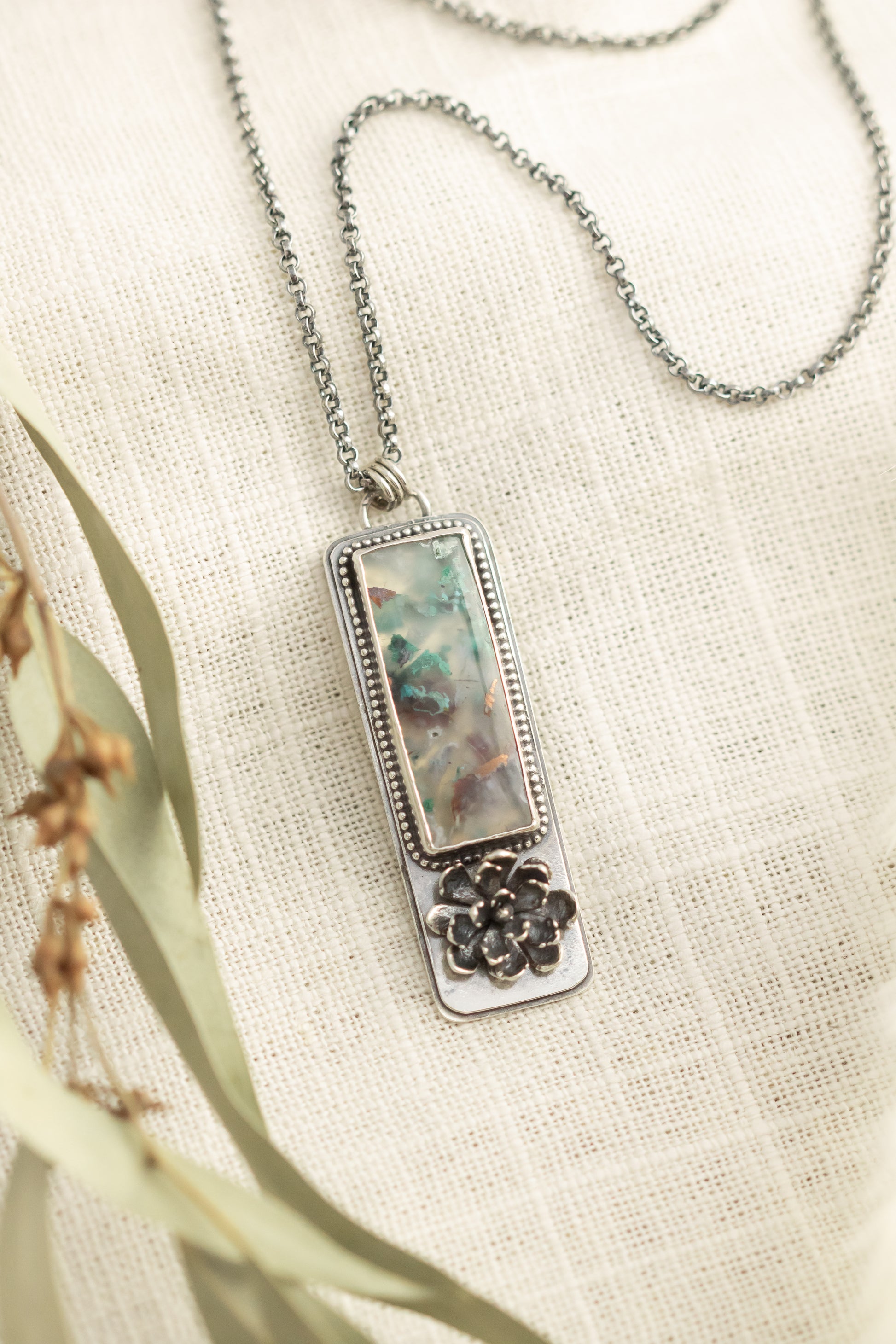 Sterling silver necklace with a sugar water flower agate pendant and succulent accent, laid flat on a beige fabric with dried eucalyptus leaves.