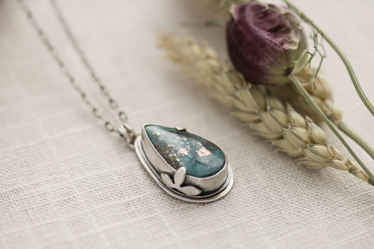 Close-up of a handmade sterling silver necklace with a teardrop-shaped Morenci 2 turquoise stone, displayed on a textured beige fabric with dried flowers and wheat stalks in the background.