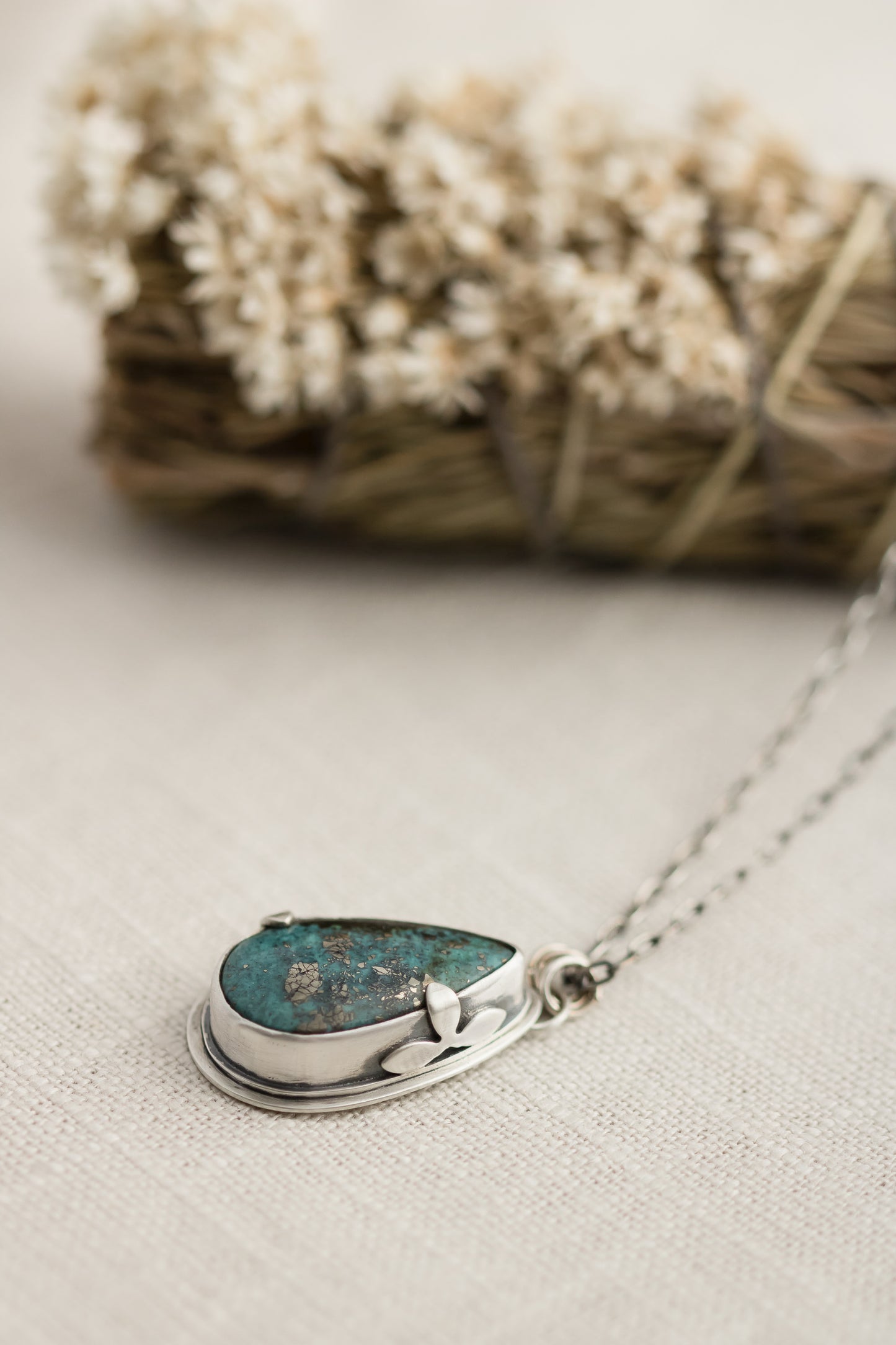Close-up of a handmade sterling silver necklace with a teardrop-shaped Morenci 2 turquoise stone, displayed on a textured beige fabric with dried flowers and wheat stalks in the background.