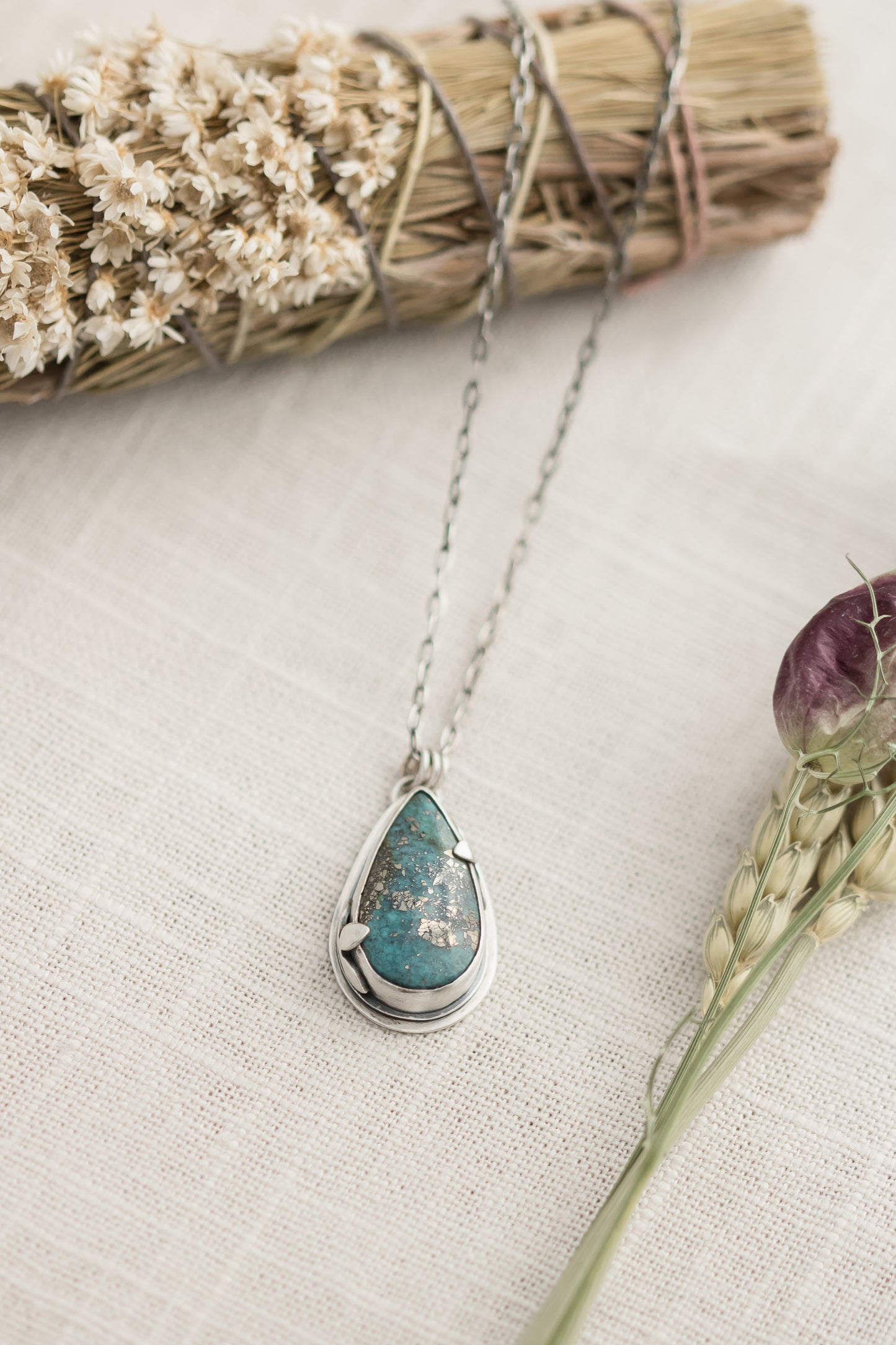 Close-up of a handmade sterling silver necklace with a teardrop-shaped Morenci 2 turquoise stone, displayed on a textured beige fabric with dried flowers and wheat stalks in the background.
