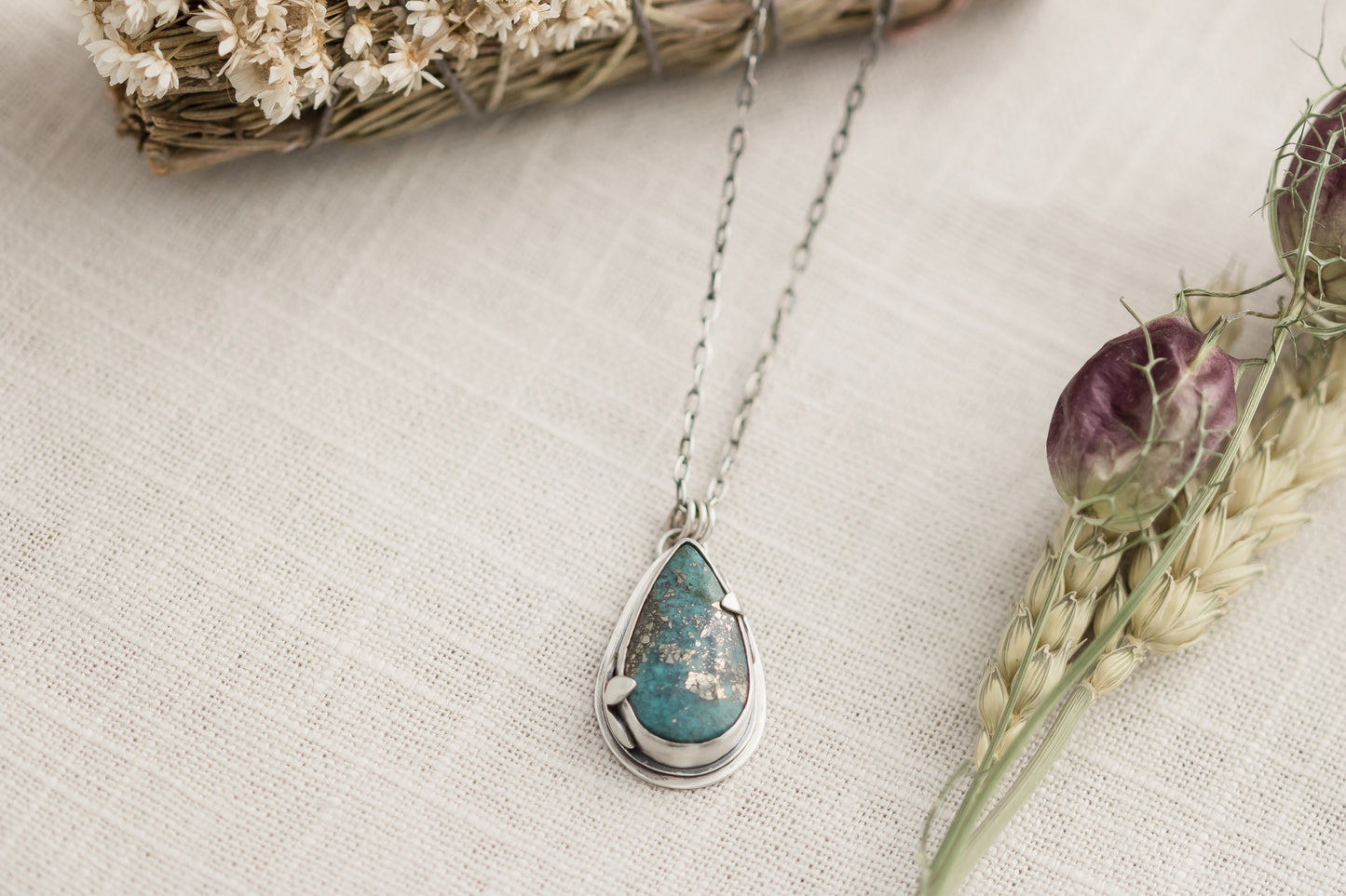 Close-up of a handmade sterling silver necklace with a teardrop-shaped Morenci 2 turquoise stone, displayed on a textured beige fabric with dried flowers and wheat stalks in the background.