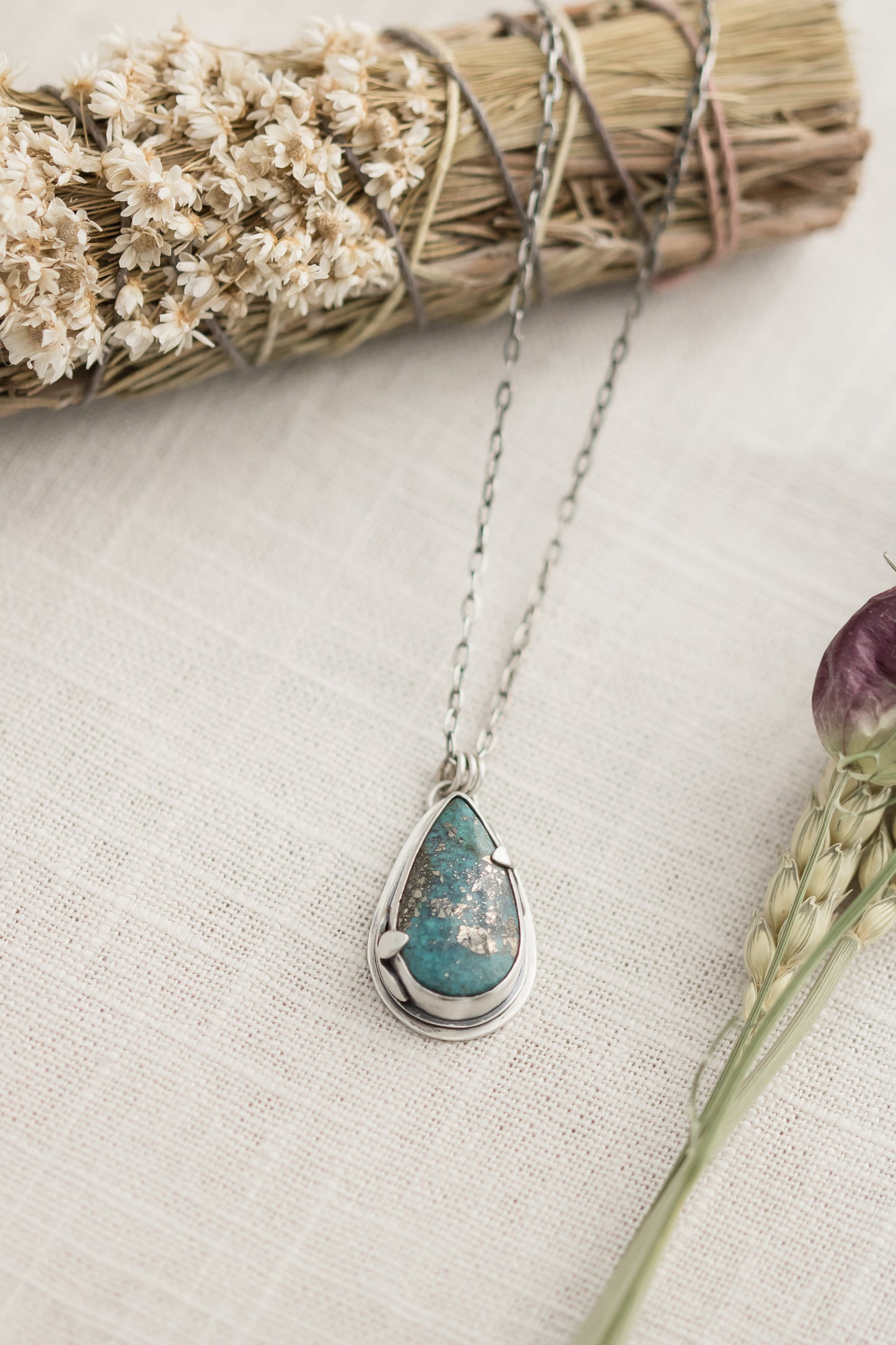 Close-up of a handmade sterling silver necklace with a teardrop-shaped Morenci 2 turquoise stone, displayed on a textured beige fabric with dried flowers and wheat stalks in the background.
