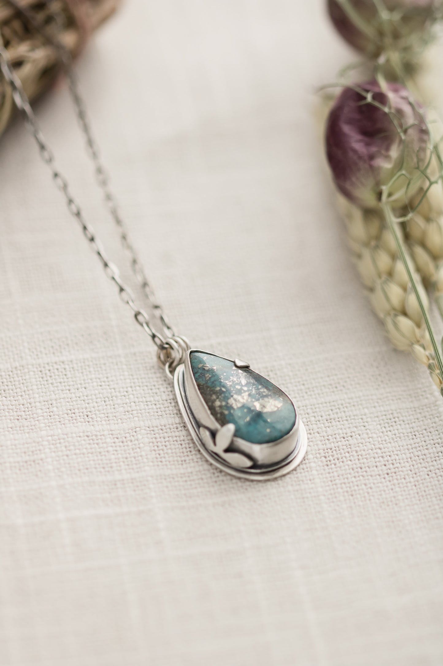 Close-up of a handmade sterling silver necklace with a teardrop-shaped Morenci 2 turquoise stone, displayed on a textured beige fabric with dried flowers and wheat stalks in the background.