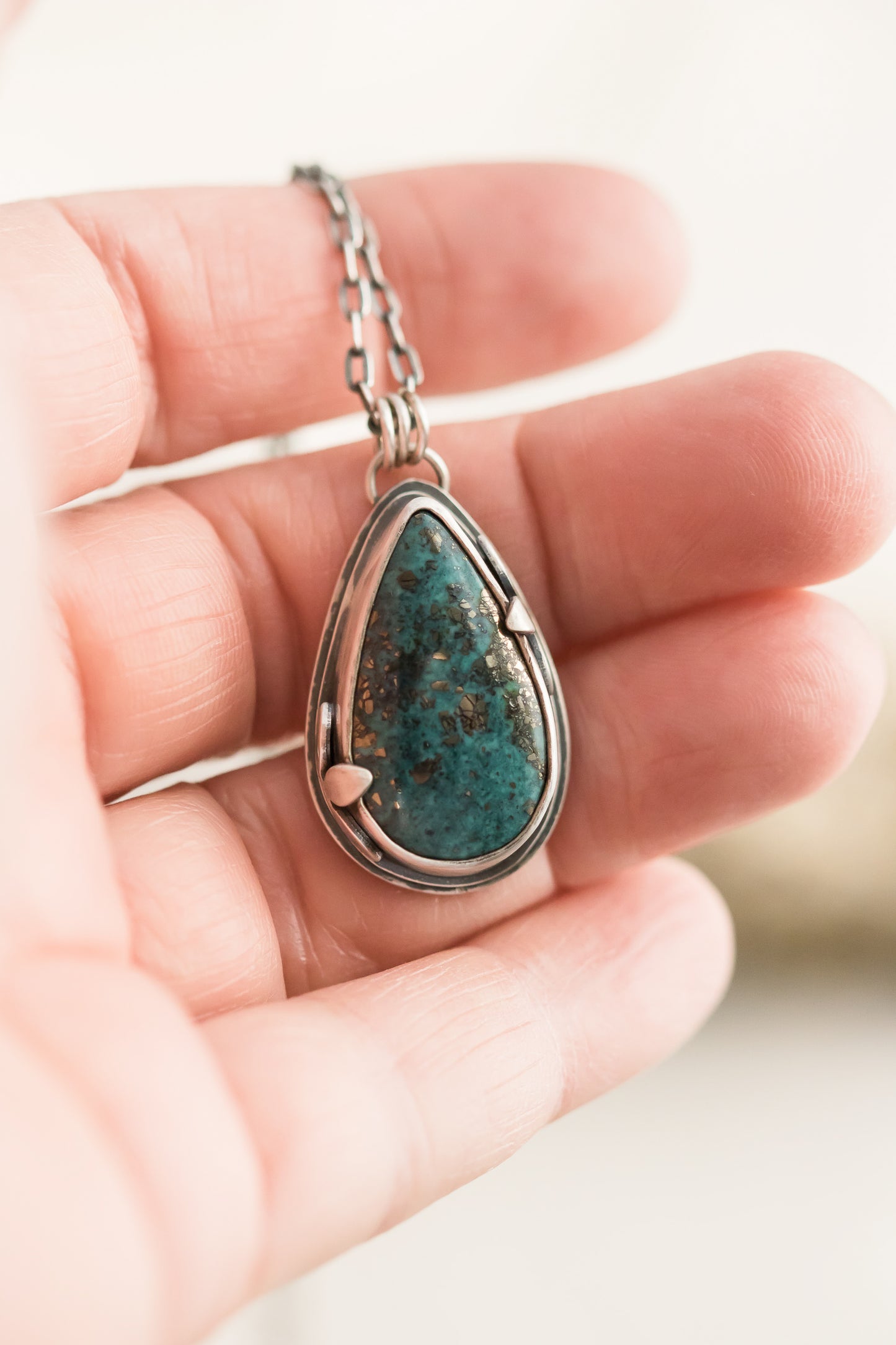 Hand holding a handmade sterling silver necklace with a Morenci 2 turquoise teardrop pendant, showcasing the vibrant stone and intricate leaf detailing against a soft background.