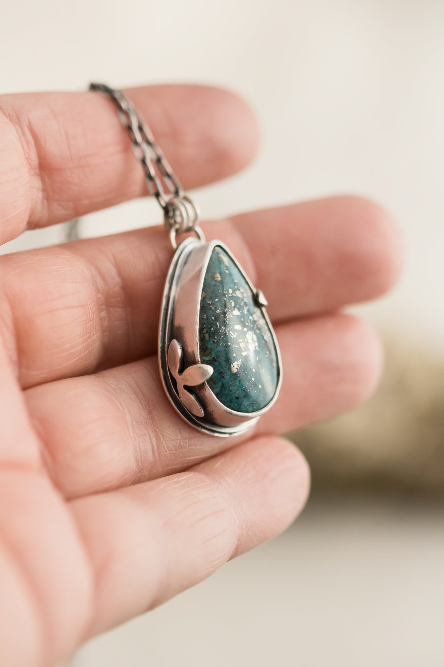 Hand holding a handmade sterling silver necklace with a Morenci 2 turquoise teardrop pendant, showcasing the vibrant stone and intricate leaf detailing against a soft background.