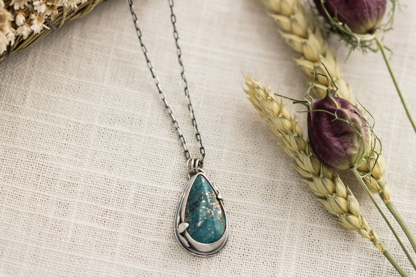 Close-up of a handmade sterling silver necklace with a teardrop-shaped Morenci 2 turquoise stone, displayed on a textured beige fabric with dried flowers and wheat stalks in the background.
