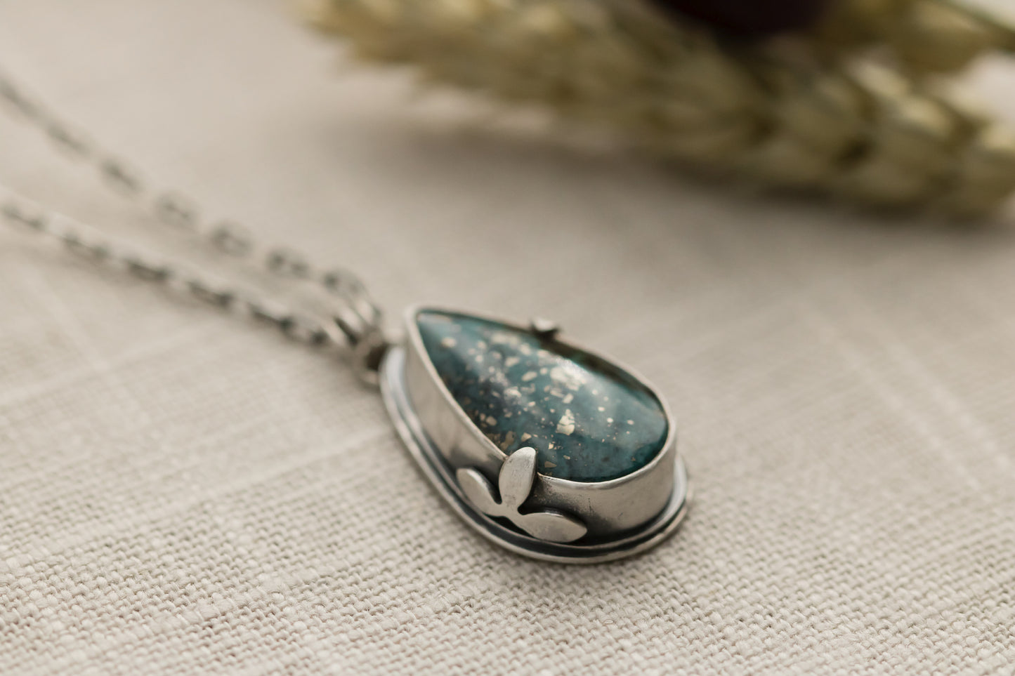 Close-up of a handmade sterling silver necklace with a teardrop-shaped Morenci 2 turquoise stone, displayed on a textured beige fabric with dried flowers and wheat stalks in the background.