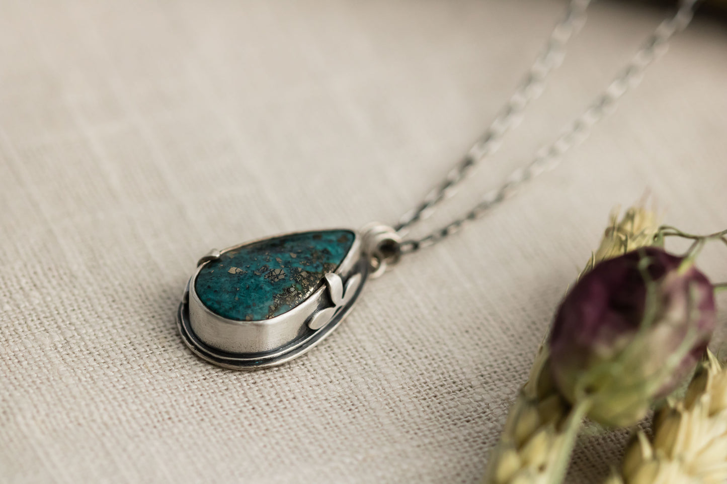 Close-up of a handmade sterling silver necklace with a teardrop-shaped Morenci 2 turquoise stone, displayed on a textured beige fabric with dried flowers and wheat stalks in the background.