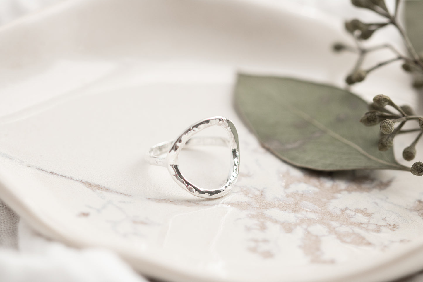 A single open circle sterling silver ring on a white marbled dish with a green leaf, set against a soft white fabric background.