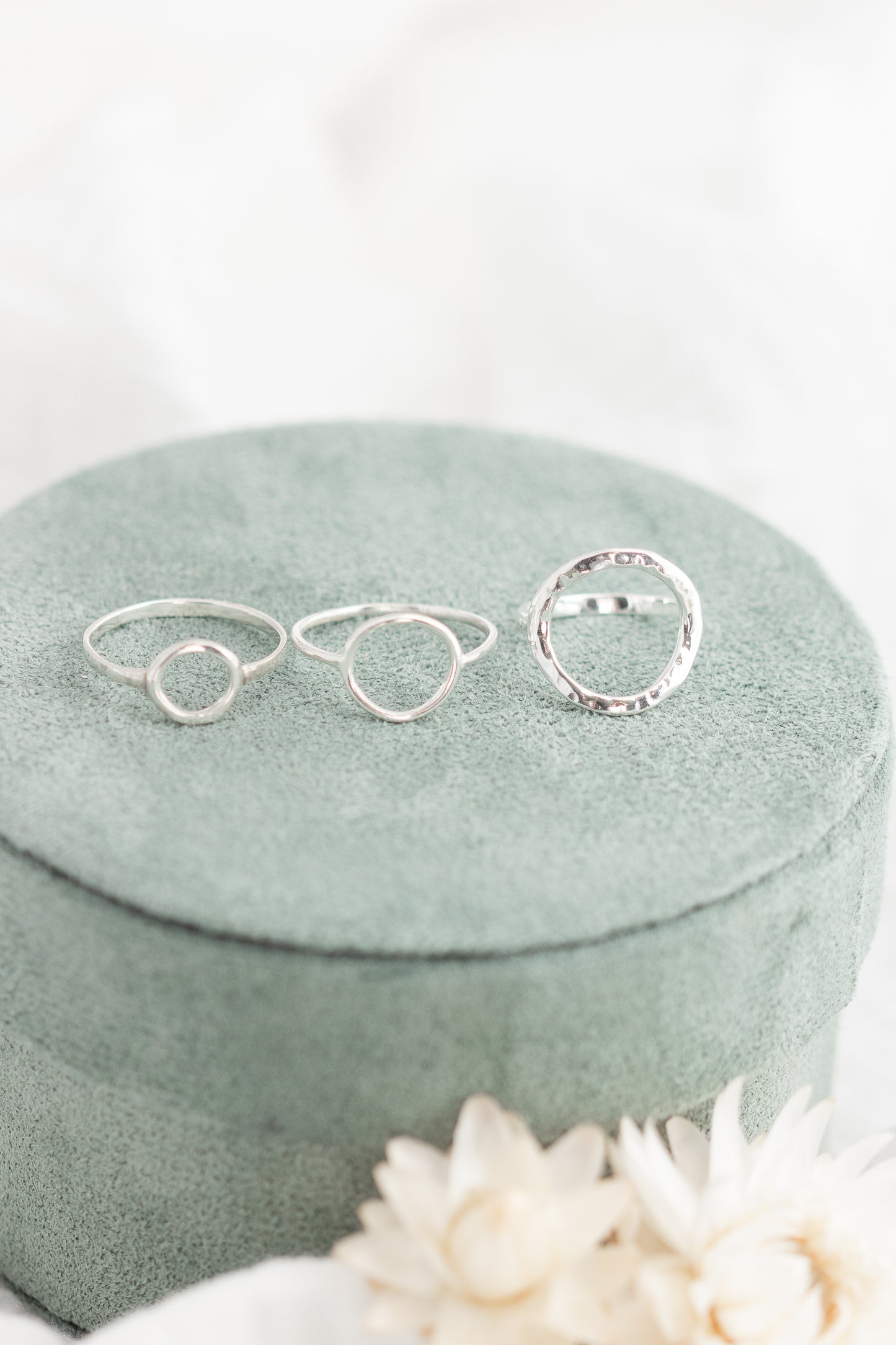 Three open circle sterling silver rings in small, medium, and large sizes, with the large ring having a hammered finish, displayed on a plush green ring box with dried flowers.