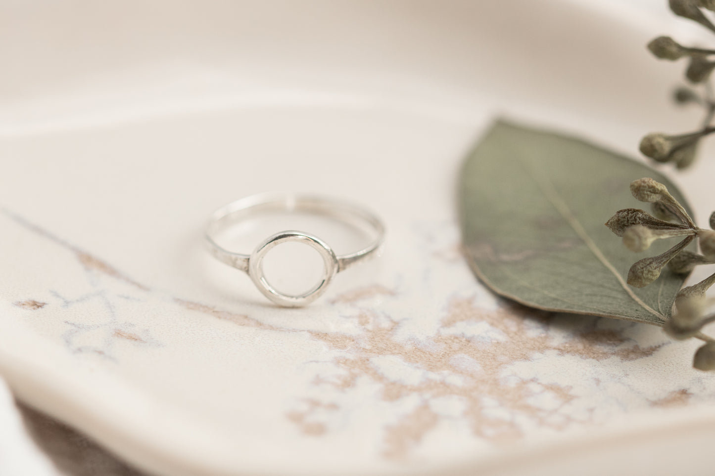A single open circle sterling silver ring on a white marbled dish with a green leaf, set against a soft white fabric background.