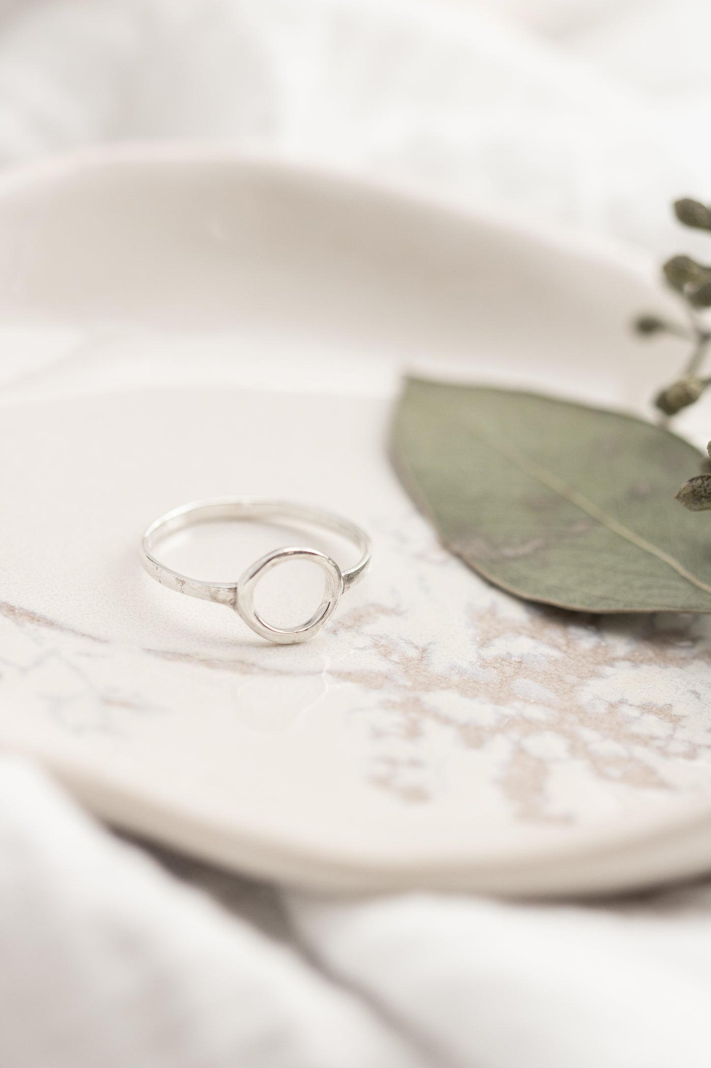 A single open circle sterling silver ring on a white marbled dish with a green leaf, set against a soft white fabric background.