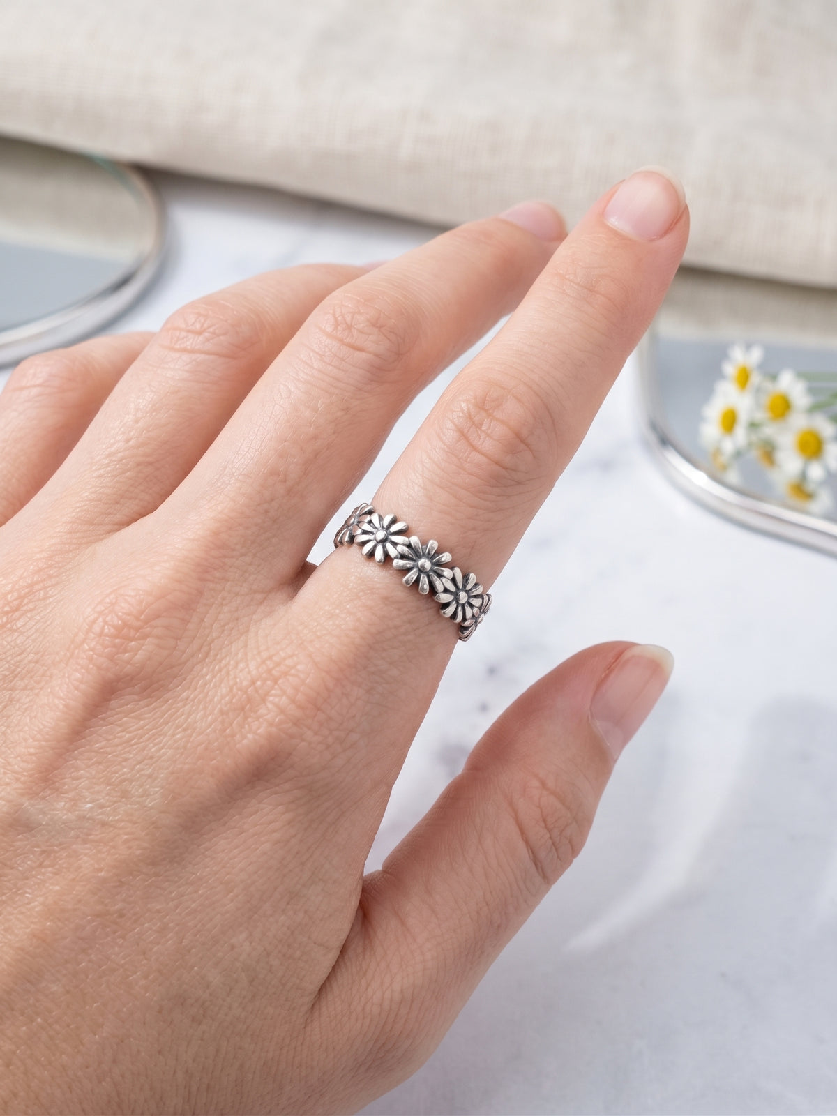 Hand modeling a dainty oxidized sterling silver daisy flower band worn on pointer finger over beige linen texture.