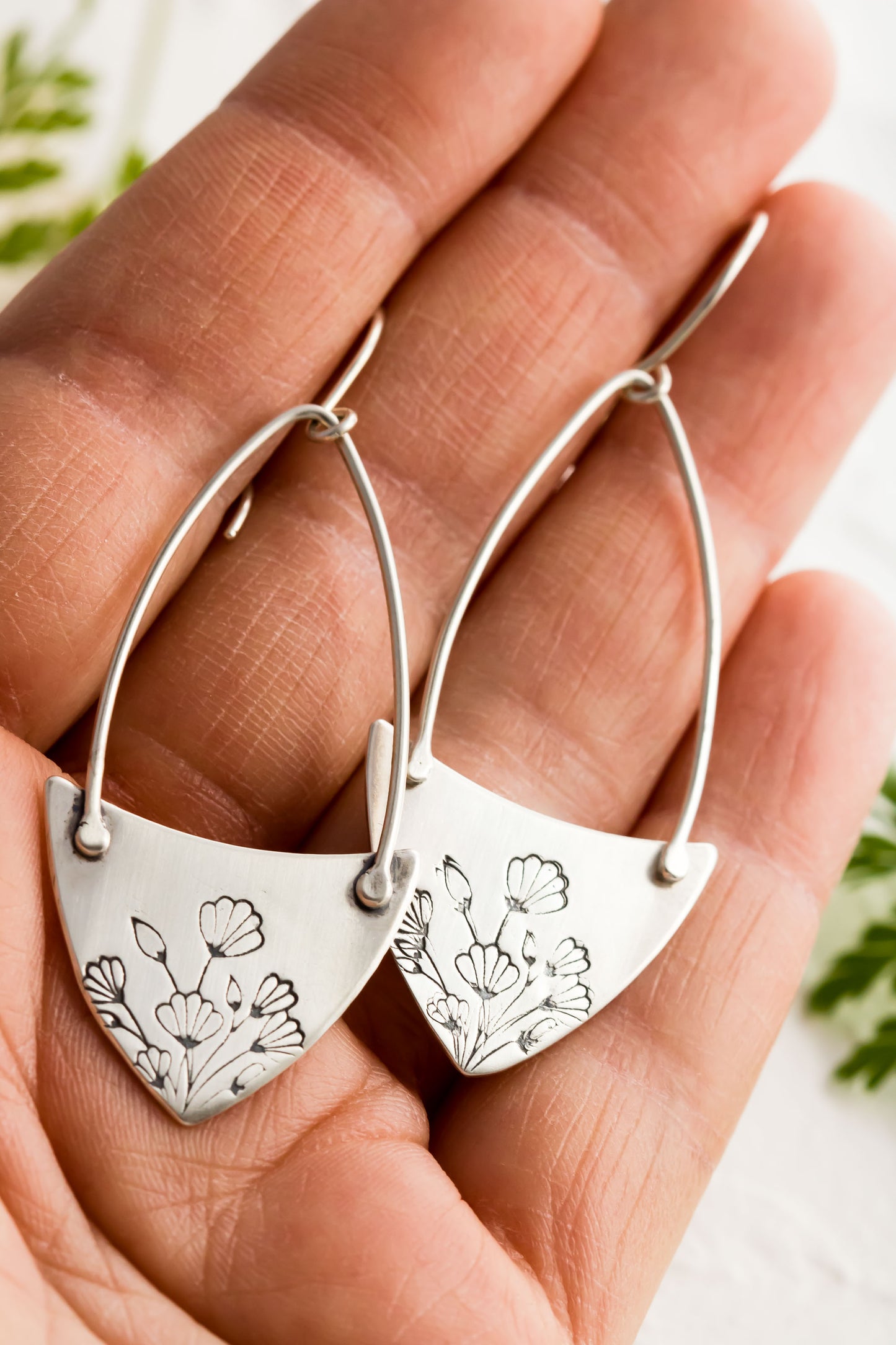 Handcrafted sterling silver Art Nouveau floral hoop earrings held I a hand with a textured background with greenery.