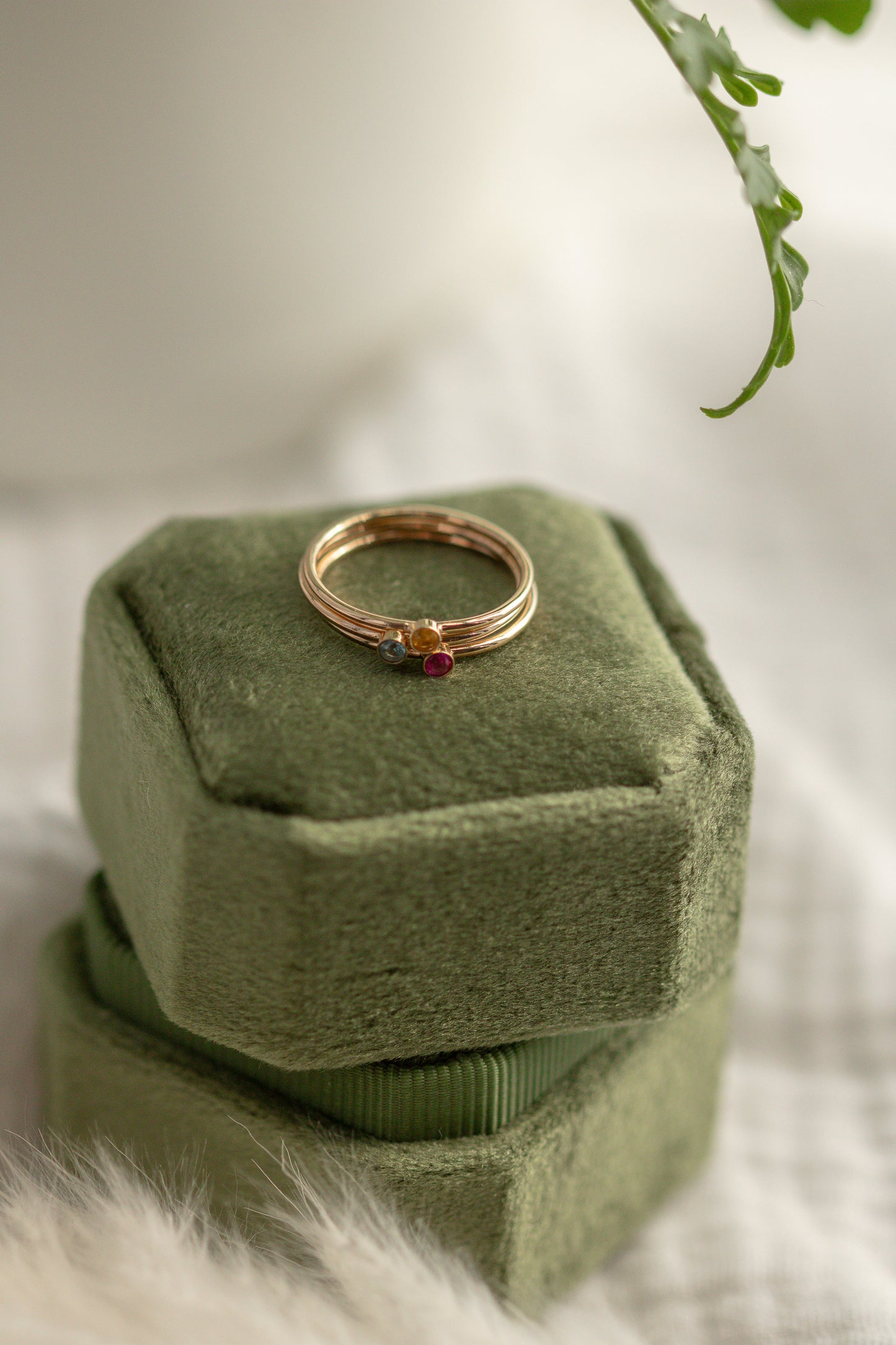 14K yellow gold birthstone stackable rings with 2mm round stones on a green velvet box, white fabric, and green plant in the background.