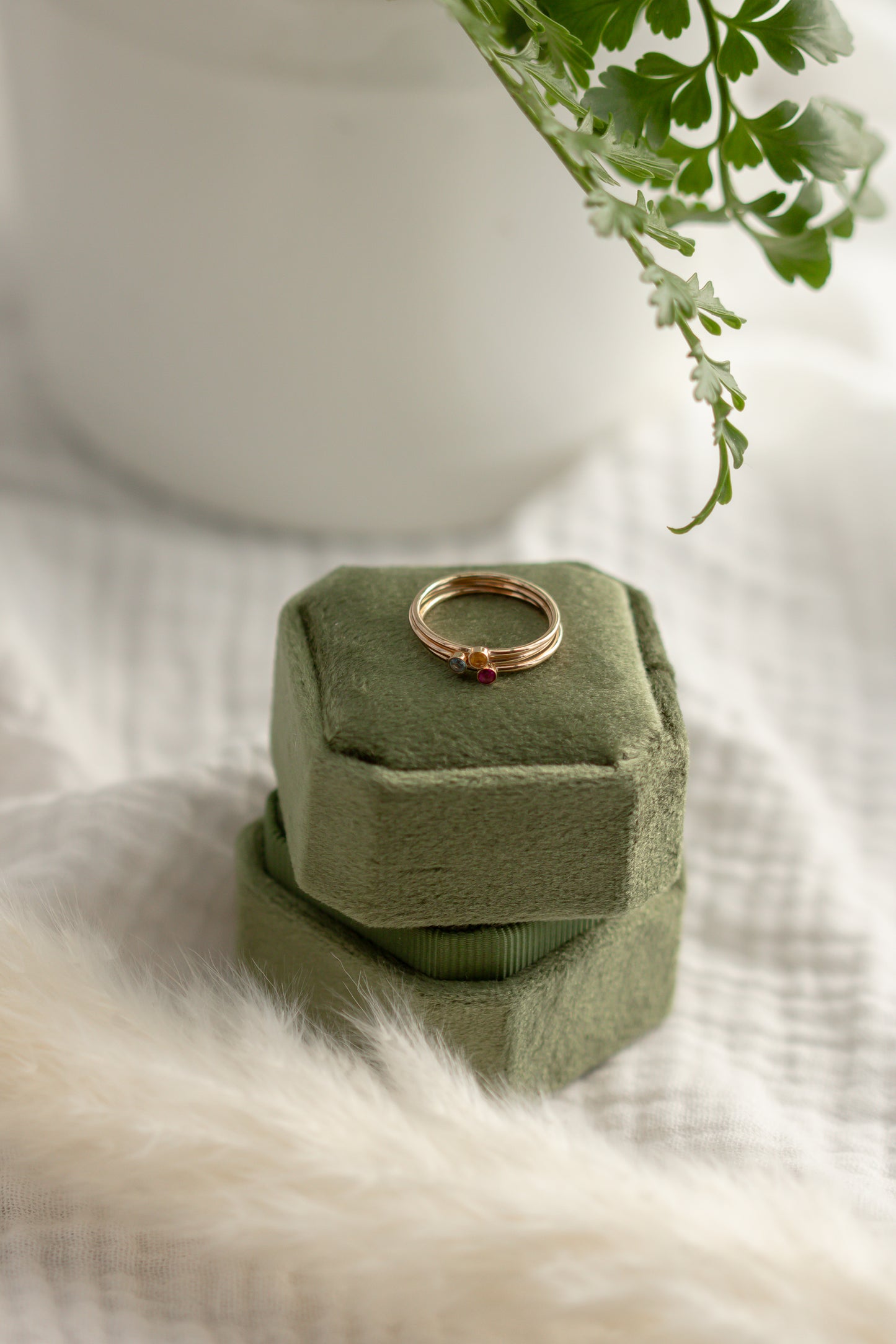 14K yellow gold birthstone stackable rings with 2mm round stones on a green velvet box, white fabric, and green plant in the background.