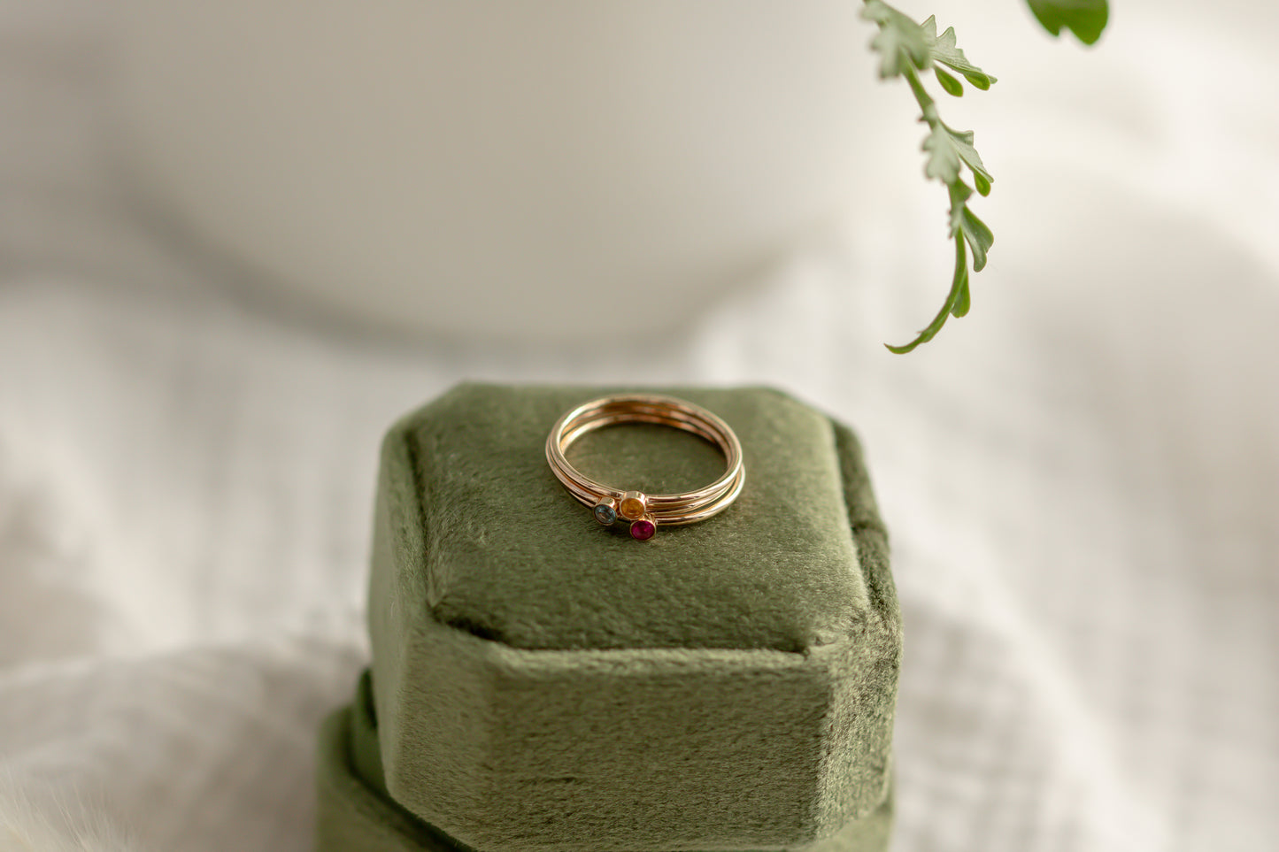 14K yellow gold birthstone stackable rings with 2mm round stones on a green velvet box, white fabric, and green plant in the background.