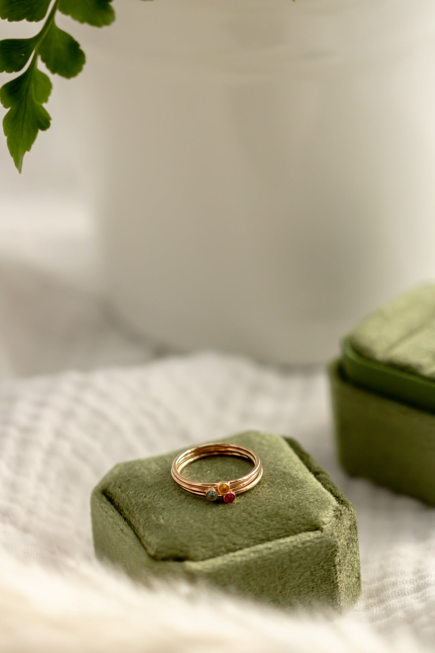 14K yellow gold birthstone stackable rings with 2mm round stones on a green velvet box, white fabric, and green plant in the background.