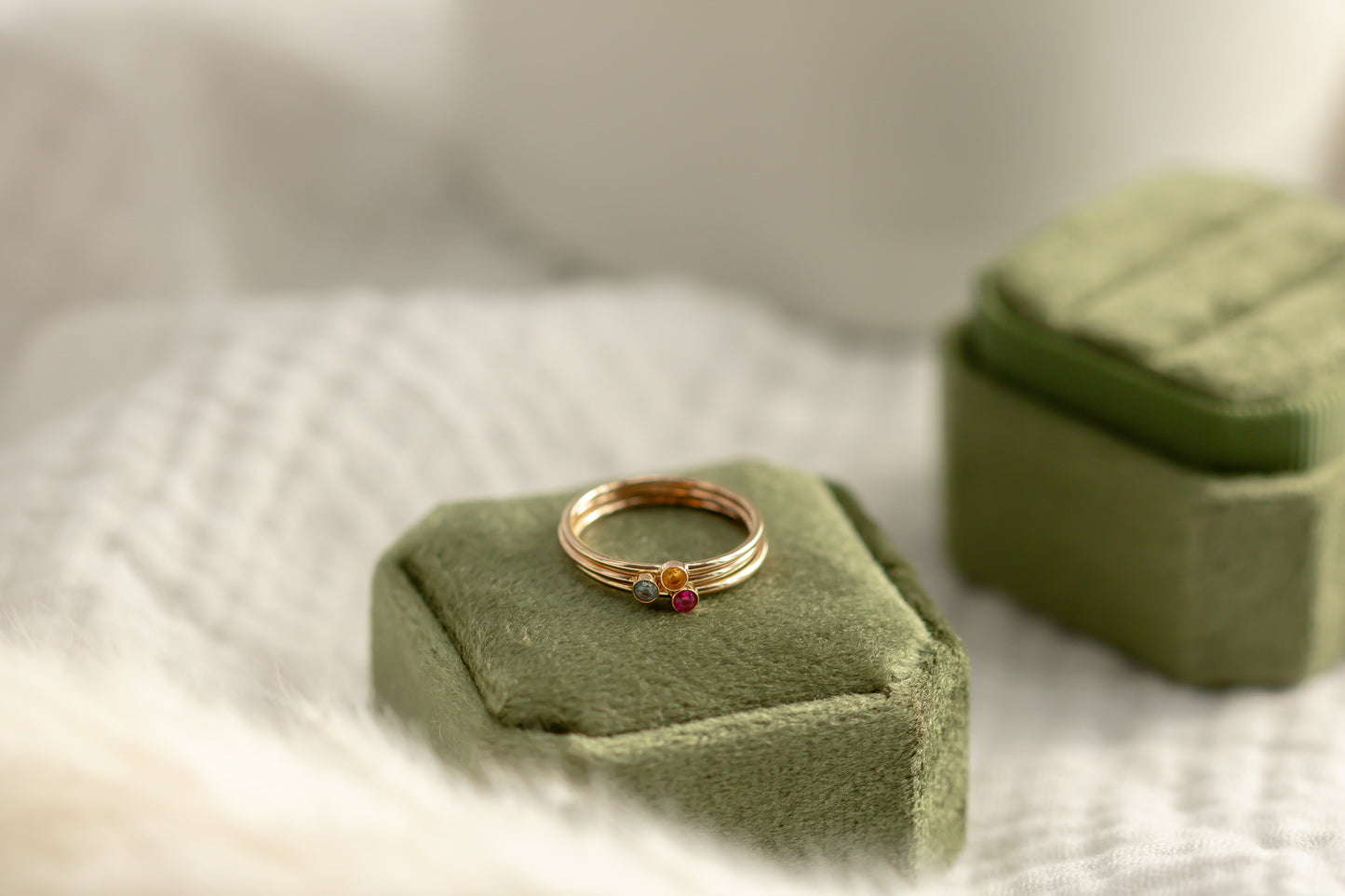 14K yellow gold birthstone stackable rings with 2mm round stones on a green velvet box, white fabric, and green plant in the background.
