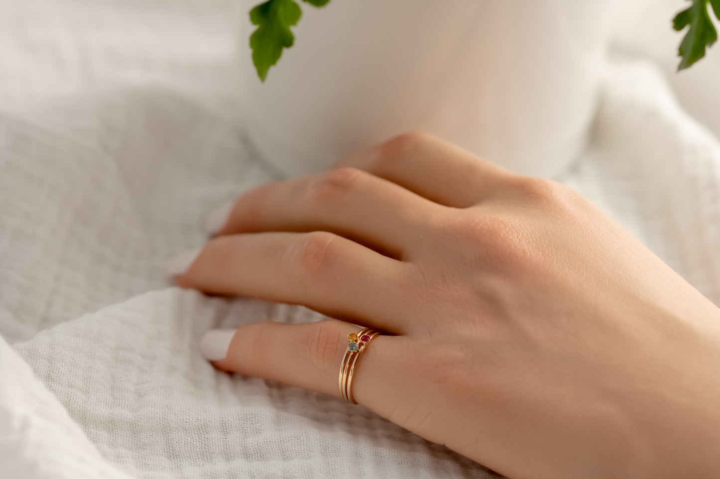 Hand wearing 14K yellow gold birthstone stackable rings with 2mm round stones, sizes 4-10, on white fabric with green plant in background.