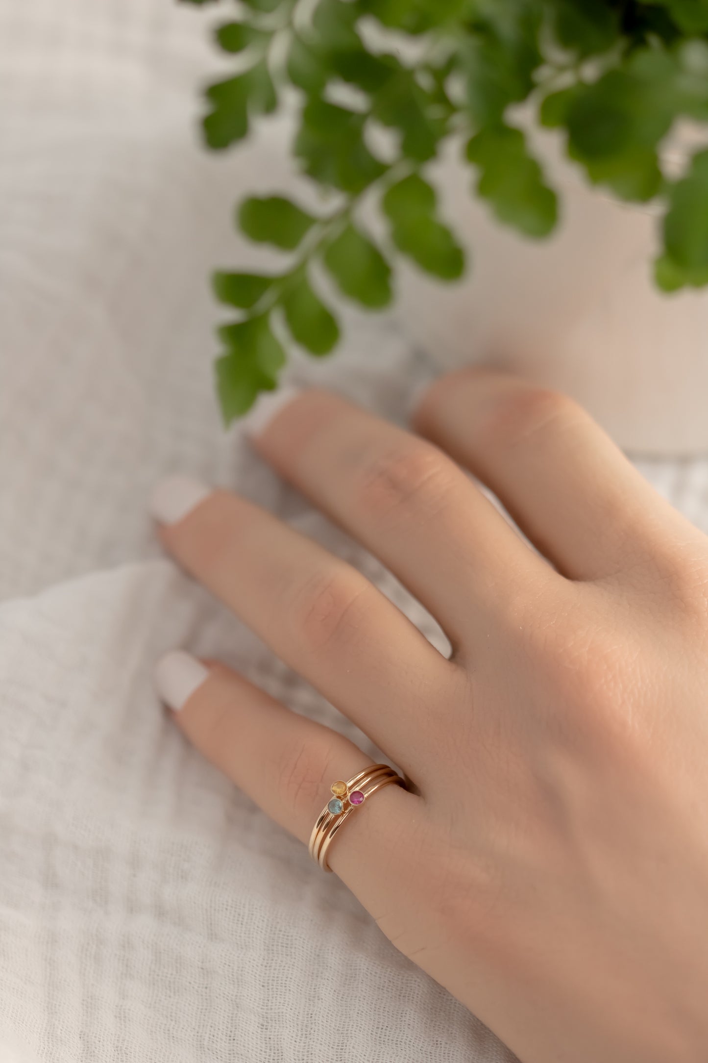 Hand wearing 14K yellow gold birthstone stackable rings with 2mm round stones, sizes 4-10, on white fabric with green plant in background.