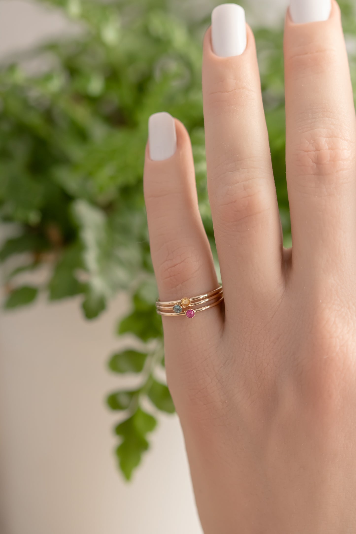 Hand wearing 14K yellow gold birthstone stackable rings with 2mm round stones, sizes 4-10, on white fabric with green plant in background.