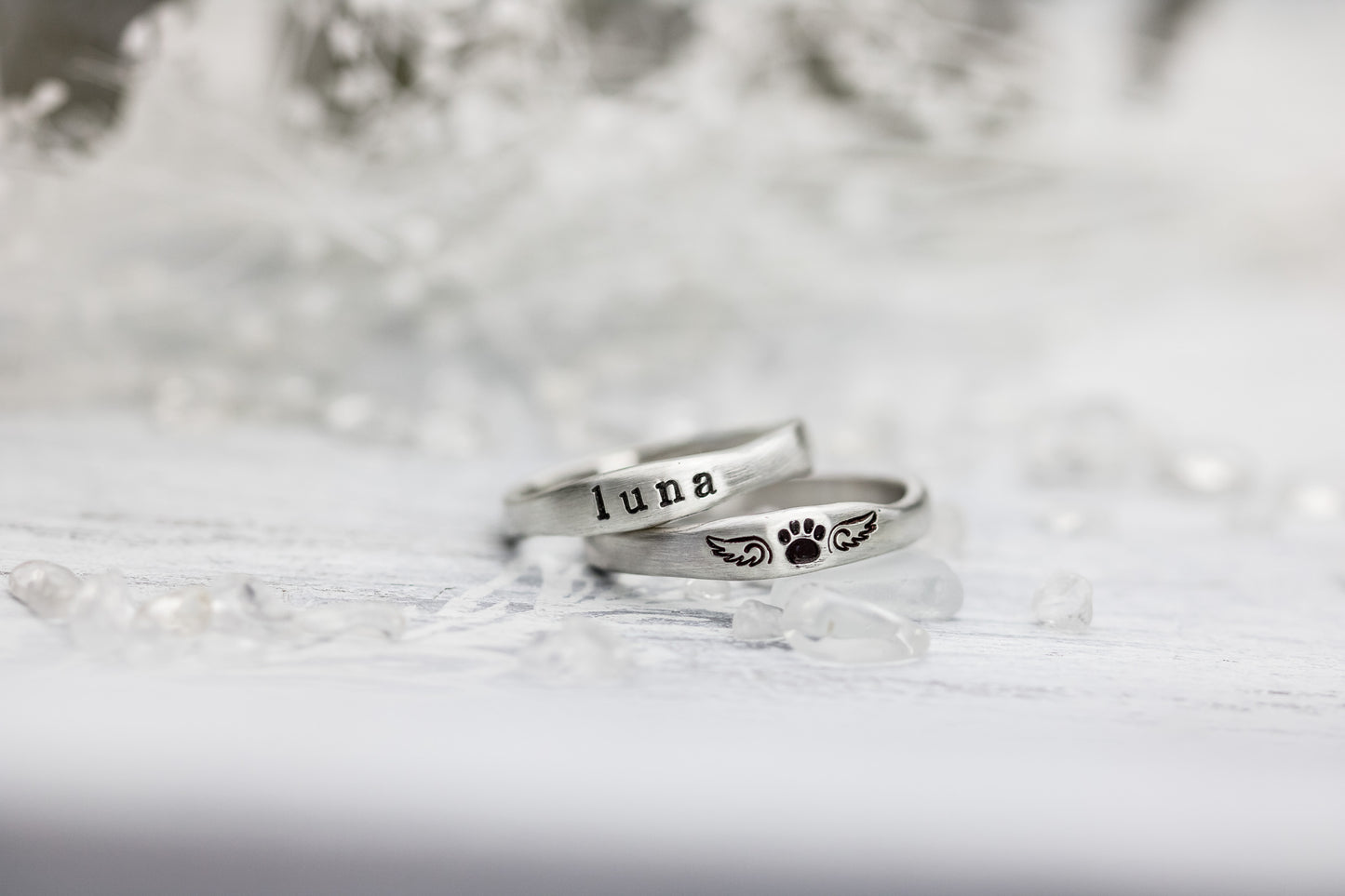Two sterling silver rings on a wooden cone, one stamped with "LUNA" and paw print with wings, against a soft white background.
