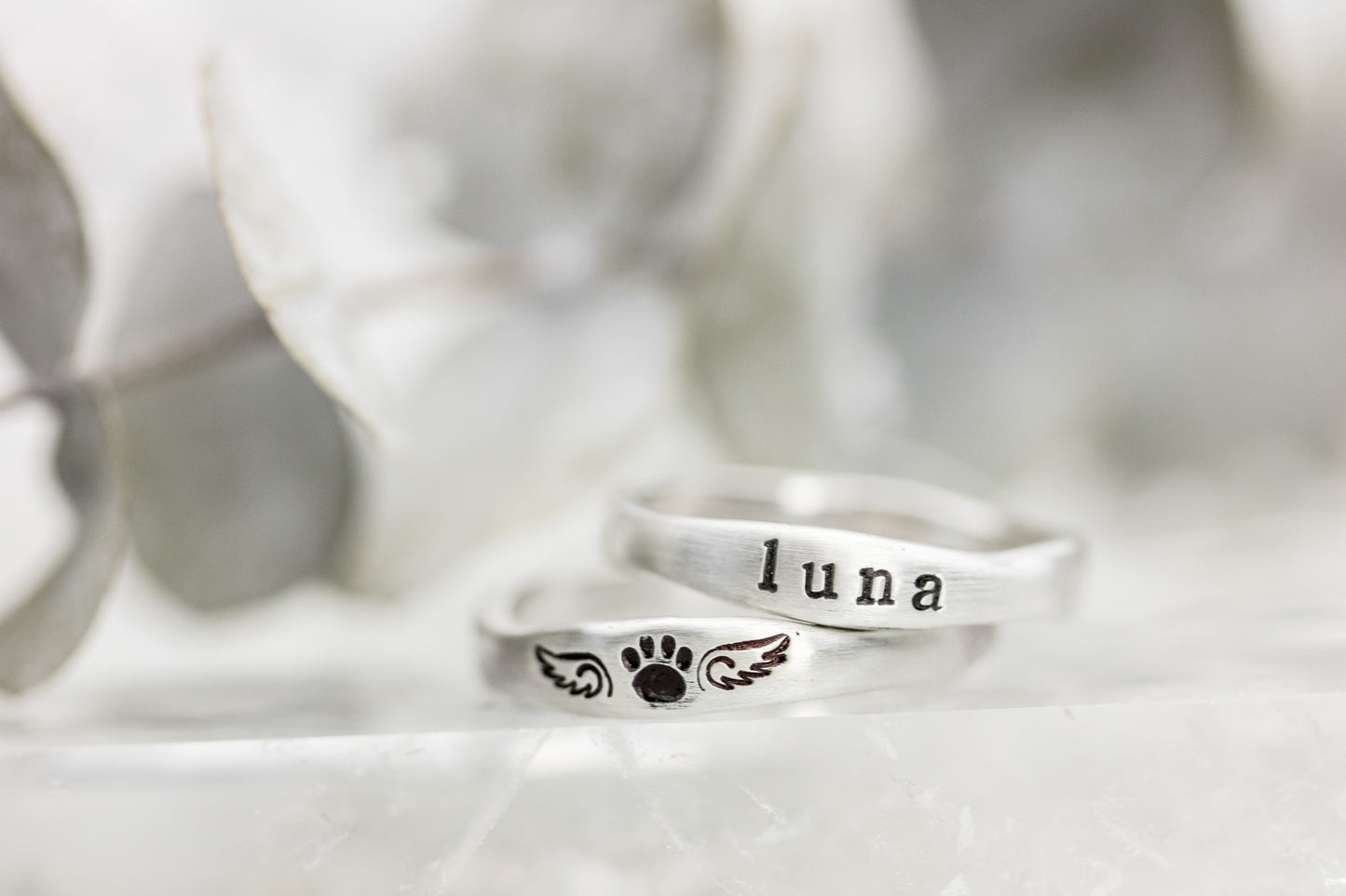 Two sterling silver rings on a wooden cone, one stamped with "LUNA" and paw print with wings, against a soft white background.