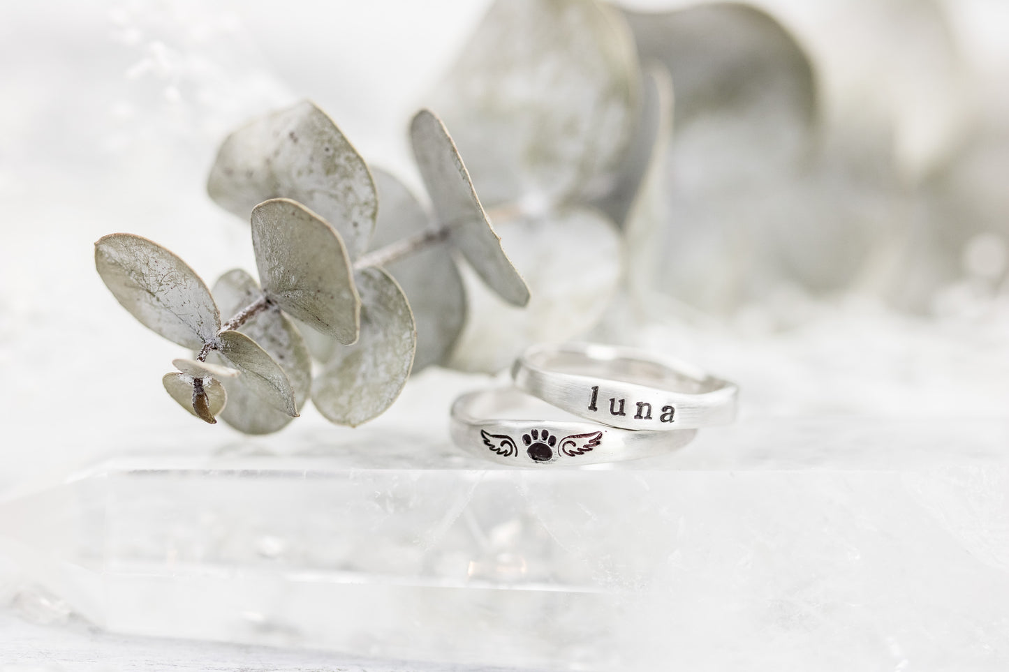 Two sterling silver rings on a wooden cone, one stamped with "LUNA" and paw print with wings, against a soft white background.