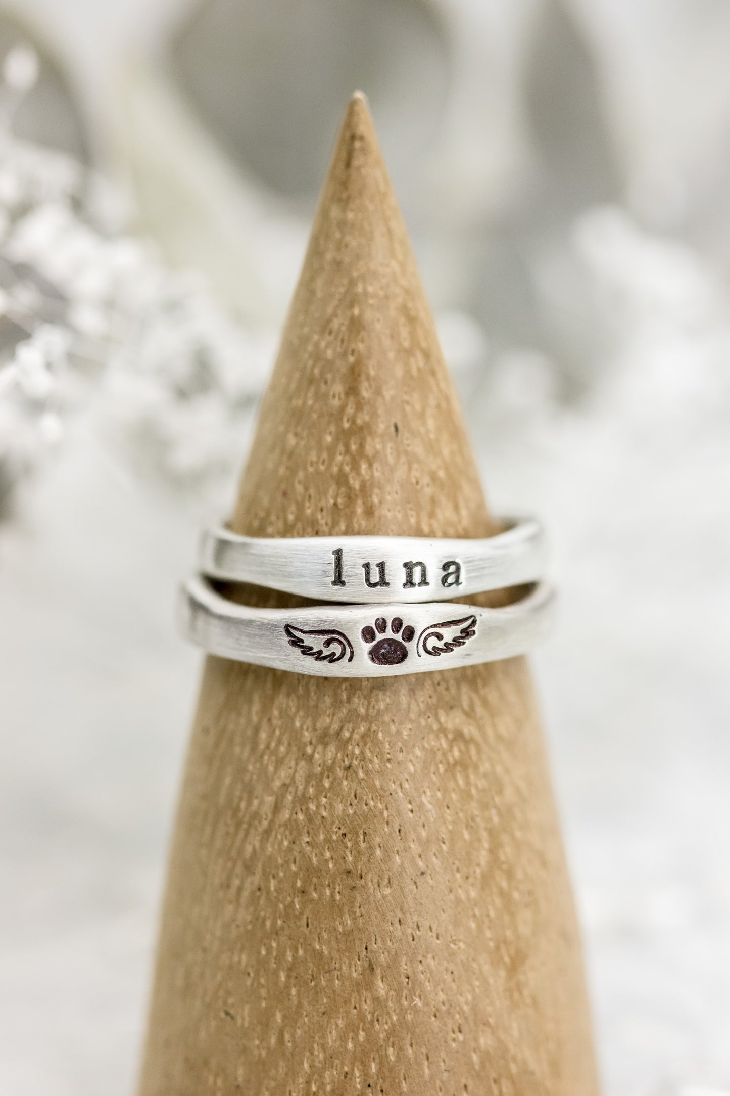 Two sterling silver rings on a wooden cone, one stamped with "LUNA" and paw print with wings, against a soft white background.