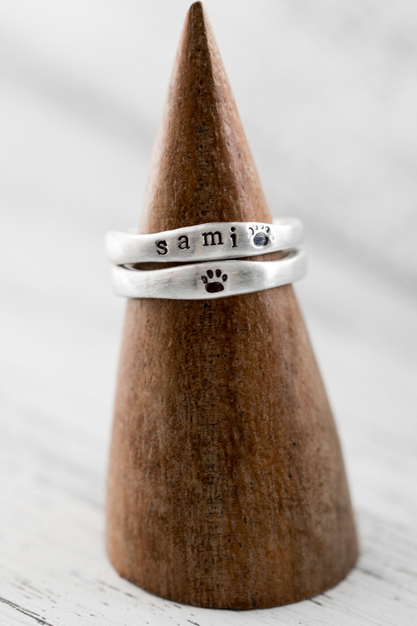 Two sterling silver rings on a wooden cone, one stamped with "SAMI" and paw prints, the other with a paw print, against a soft white background.