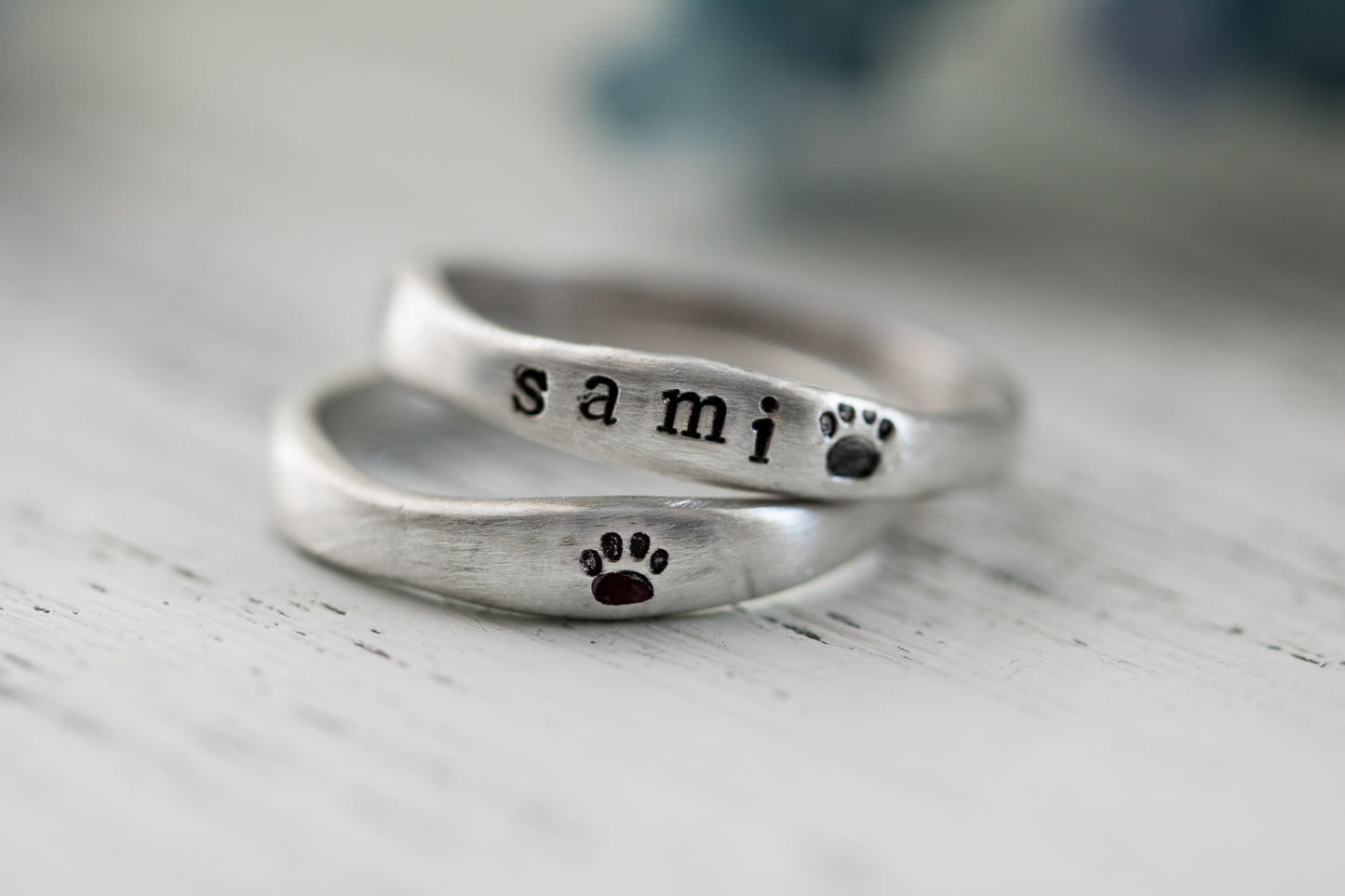 Close-up of two sterling silver rings with "SAMI" and paw prints hand-stamped, placed on a white textured surface with a blurred background.