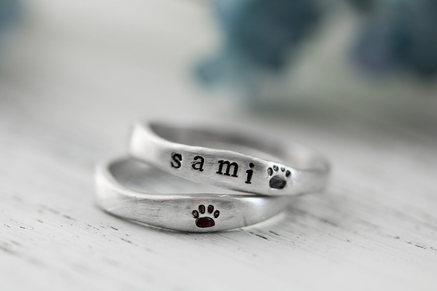 Close-up of two sterling silver rings with "SAMI" and paw prints hand-stamped, placed on a white textured surface with a blurred background.