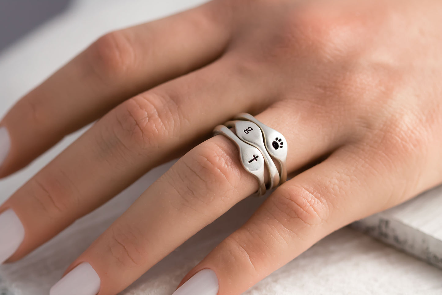 Hand with three sterling silver rings stamped with a paw print, cross, and "a" on a white textured background with dried flowers.