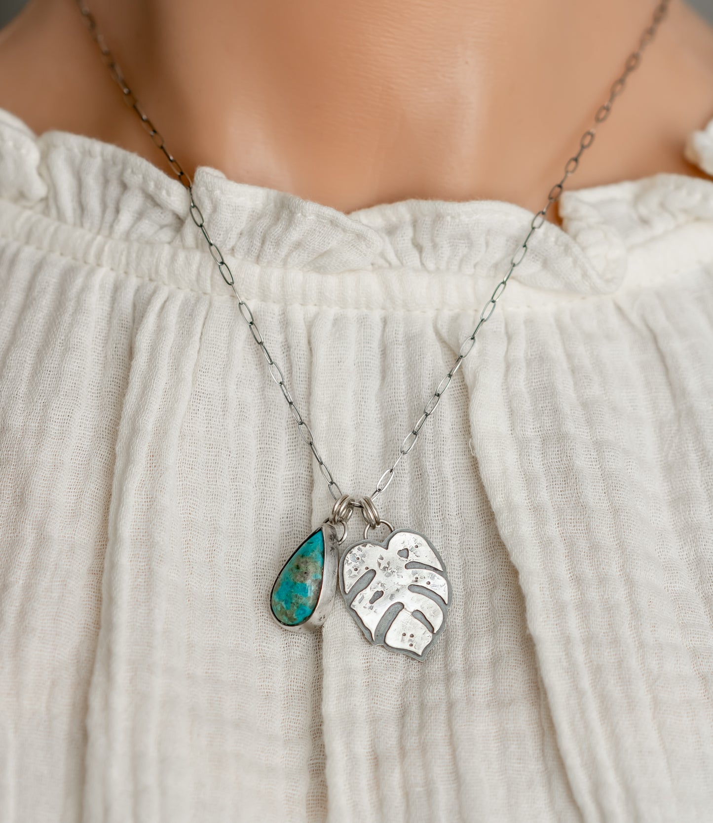 A person wears a turquoise and sterling silver Monstera leaf charm necklace on an oxidized elongated oval chain, paired with a white ruffled blouse, highlighting the boho style.