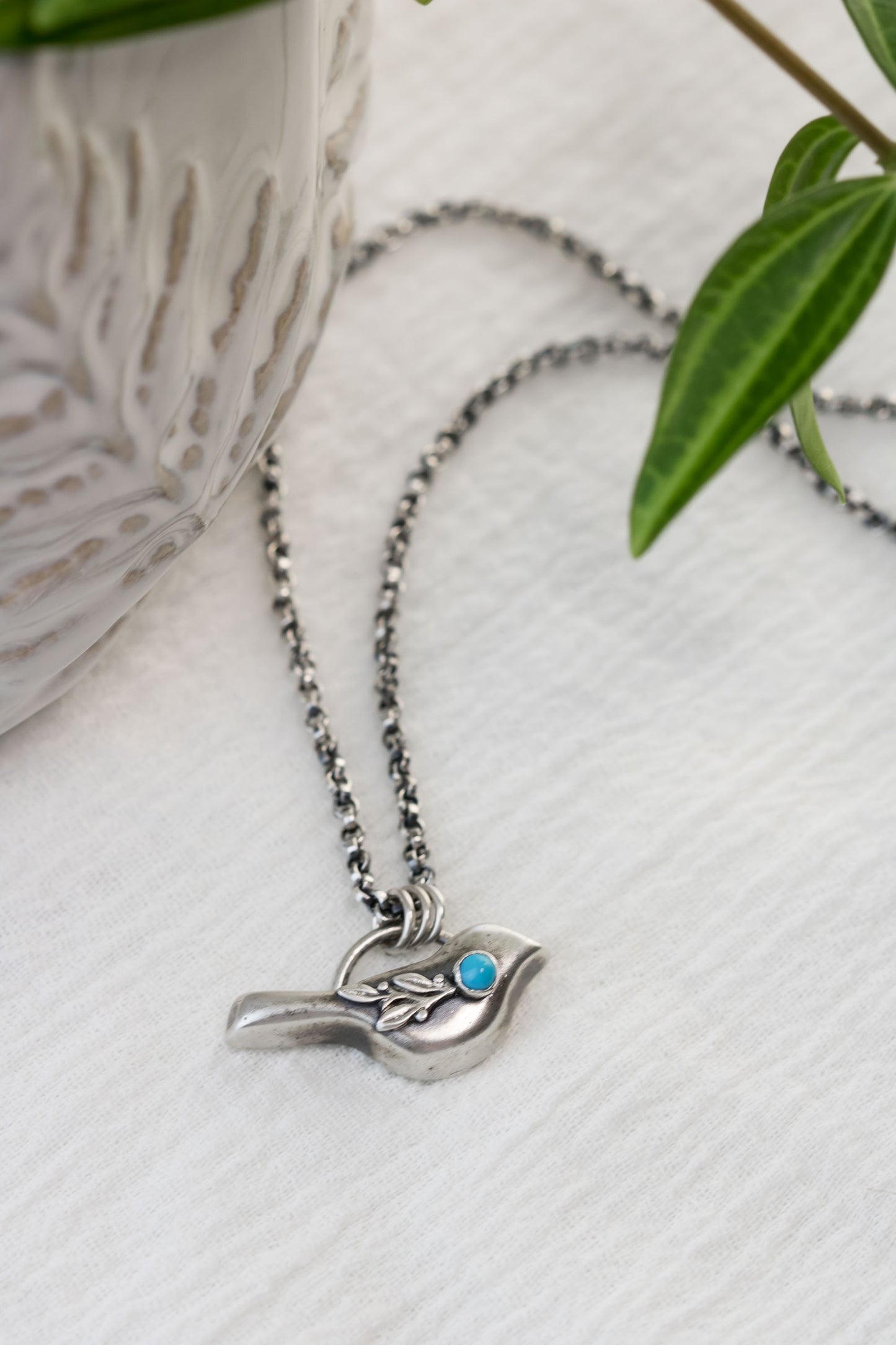 A sterling silver bird necklace with a turquoise gemstone accent, hanging on a rolo chain, displayed on a white textured surface with green leaves and a decorative bowl in the background.