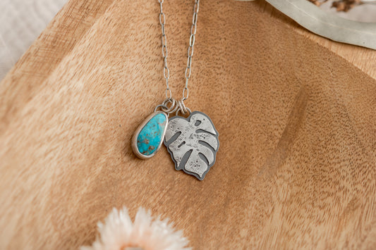 A turquoise and sterling silver Monstera leaf charm necklace on an oxidized elongated oval chain lies on a wooden surface, featuring 1" and 3/4" charms with a soft, natural background.