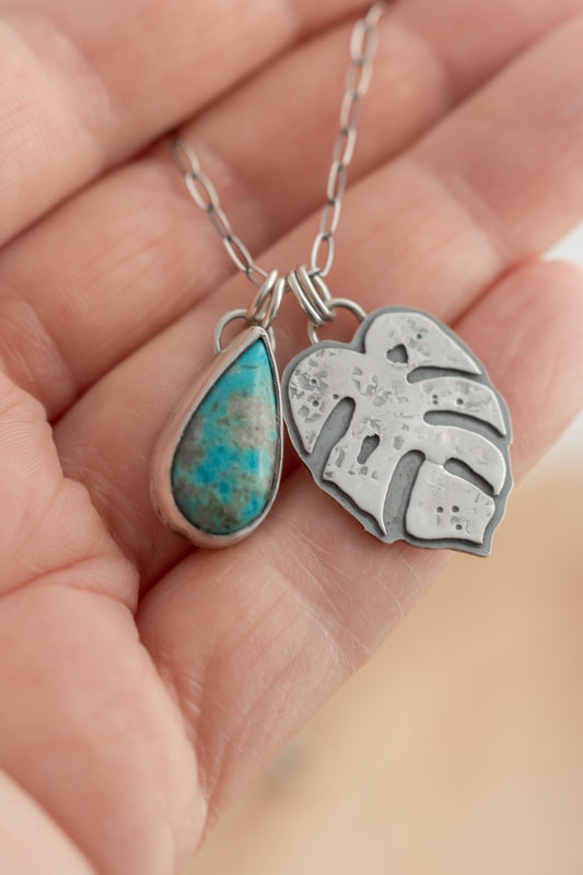 A hand holds a turquoise and sterling silver Monstera leaf charm necklace on an oxidized elongated oval chain, showcasing the 1" and 3/4" turquoise and leaf charms against a blurred background.