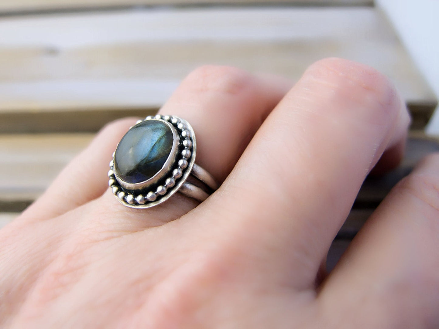 A hand wearing a sterling silver ring with a large, oval, iridescent labradorite gemstone. The gemstone is surrounded by a beaded border.