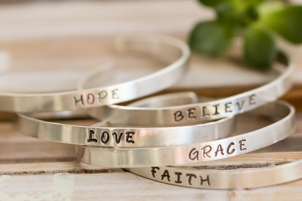 Five sterling silver bracelets with words HOPE, LOVE, GRACE, FAITH, BELIEVE, on a wood surface with a green succulent in the background