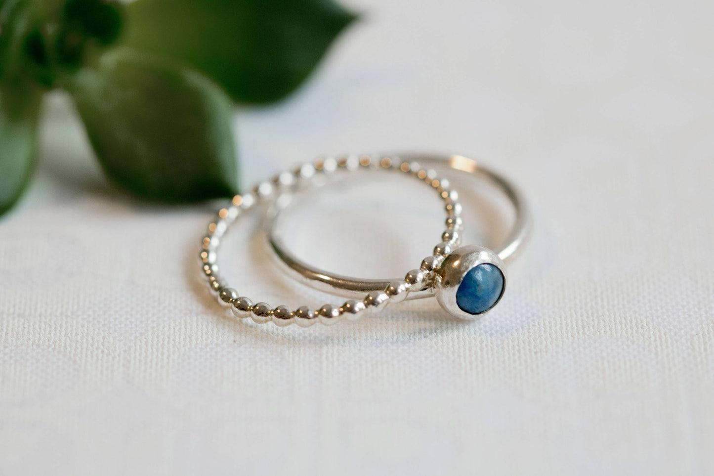 Close-up of a denim lapis ring with a beaded silver band on a white background with green leaves.