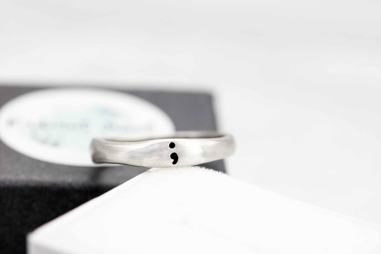 A close-up of a sterling silver semicolon awareness ring with a subtle semicolon symbol, displayed on a soft white surface with a branded box in the background, symbolizing mental health support and hope.