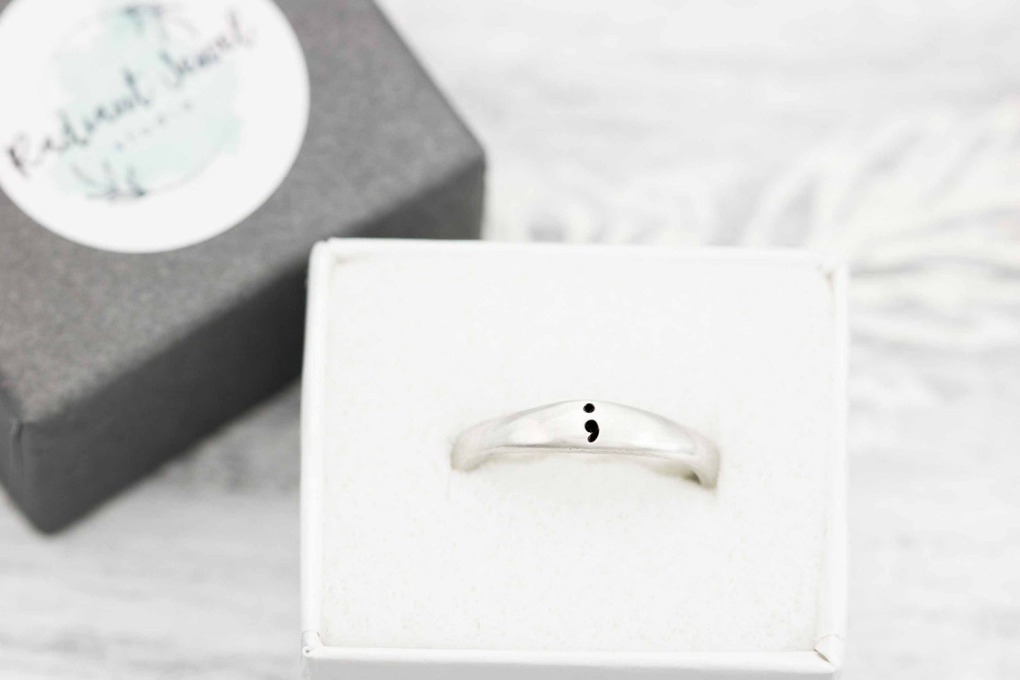A close-up of a sterling silver semicolon awareness ring with a subtle semicolon symbol, displayed on a soft white surface with a branded box in the background, symbolizing mental health support and hope.