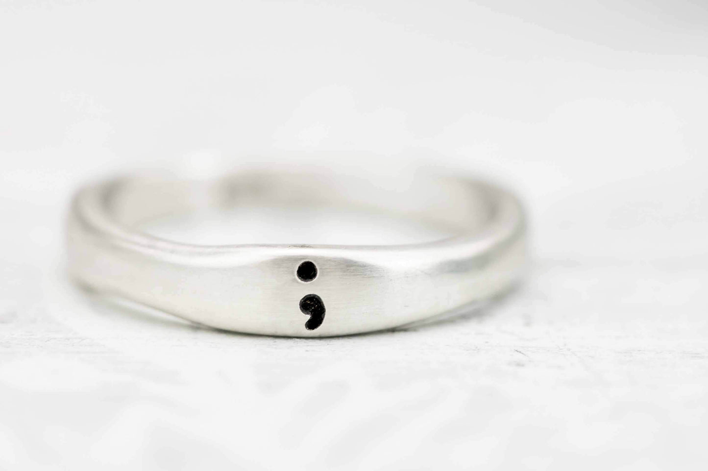 A close-up of a sterling silver semicolon awareness ring with a subtle semicolon symbol, displayed on a soft white surface symbolizing mental health support and hope.