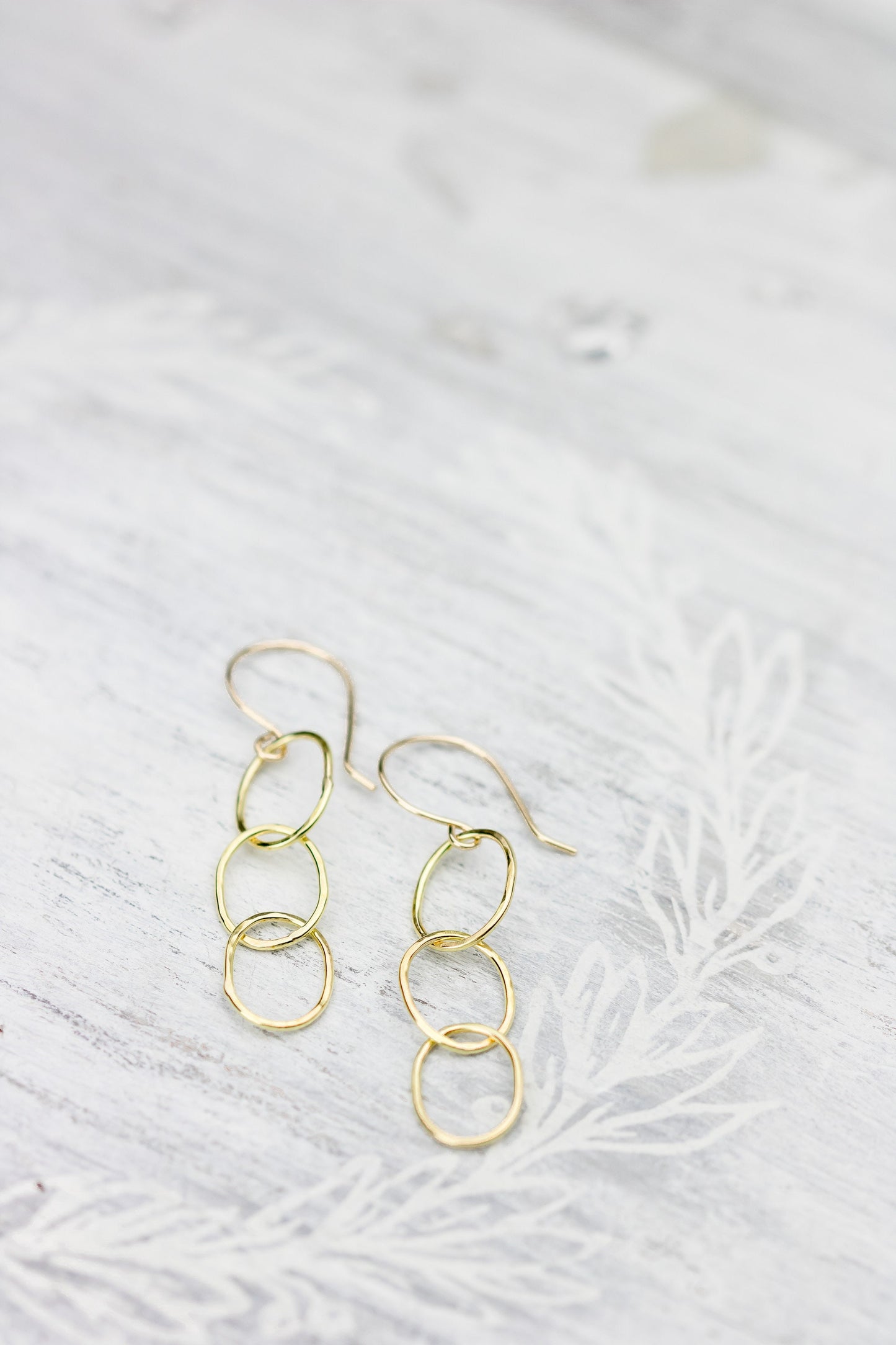 Two 18K yellow gold hammered triple hoop earrings placed on a white textured surface with a faint floral pattern, highlighting their design.
