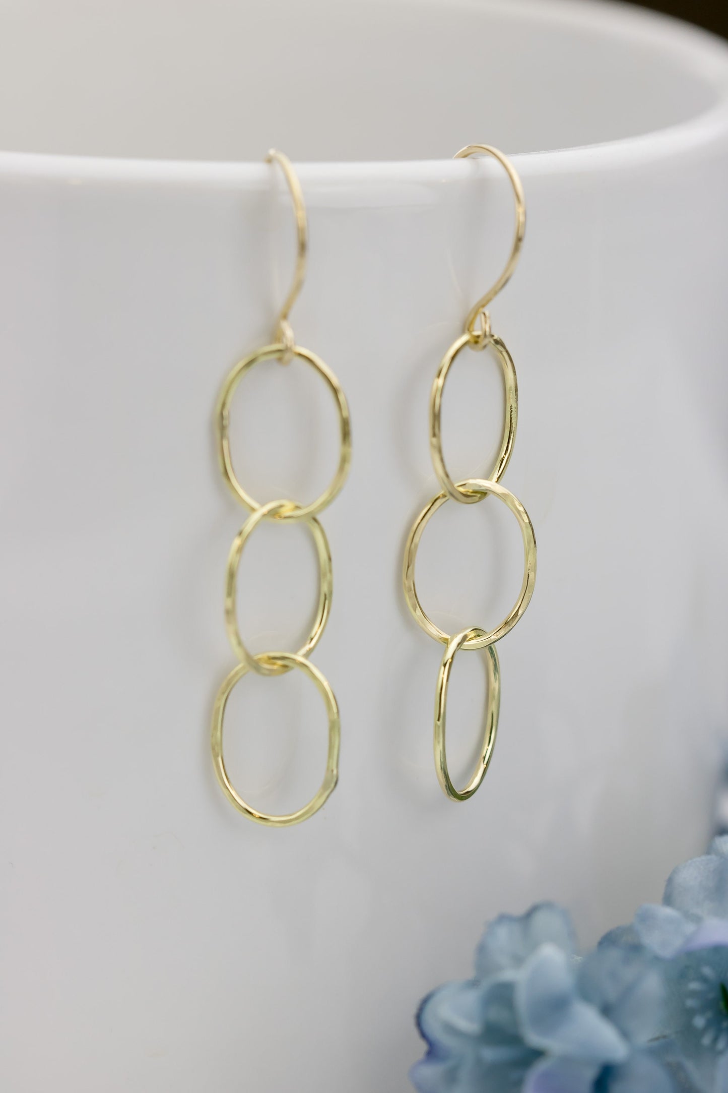 A pair of 18K yellow gold hammered triple hoop earrings draped over a white ceramic cup, with soft blue flowers in the background, showcasing their elegance.
